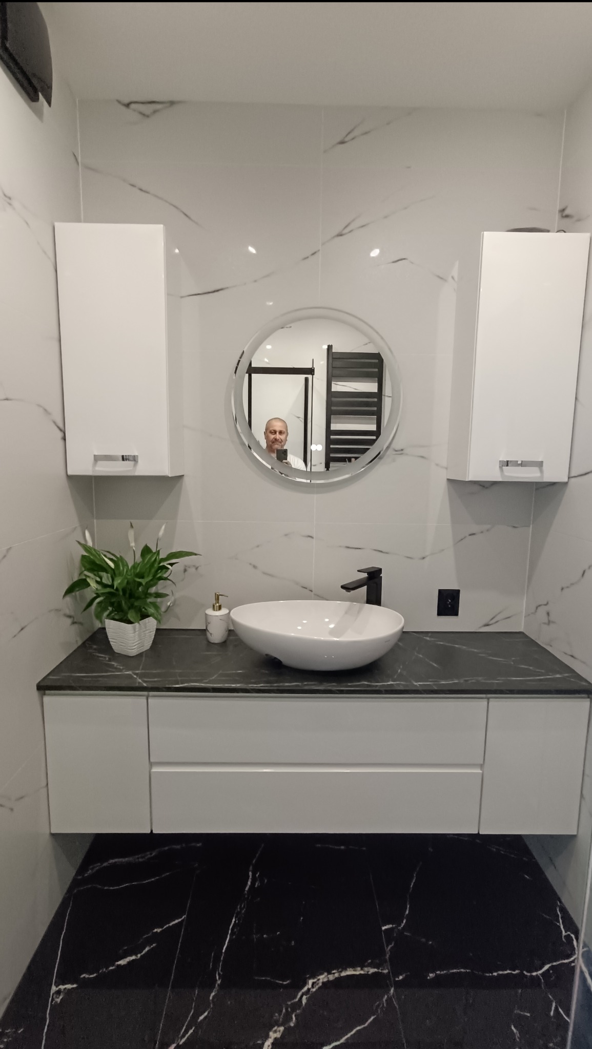 Elegancka łazienka z biało-czarnymi marmurkowymi płytkami, okrągłym lustrem z podświetleniem i białymi szafkami. Na blacie umywalka i roślina.