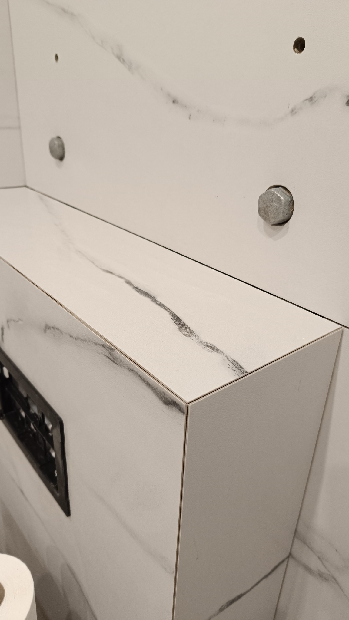 Biała zabudowa WC z płytek imitujących marmur, widoczne precyzyjne łączenia płytek, otwory w ścianie przygotowane pod montaż. Minimalistyczny design.