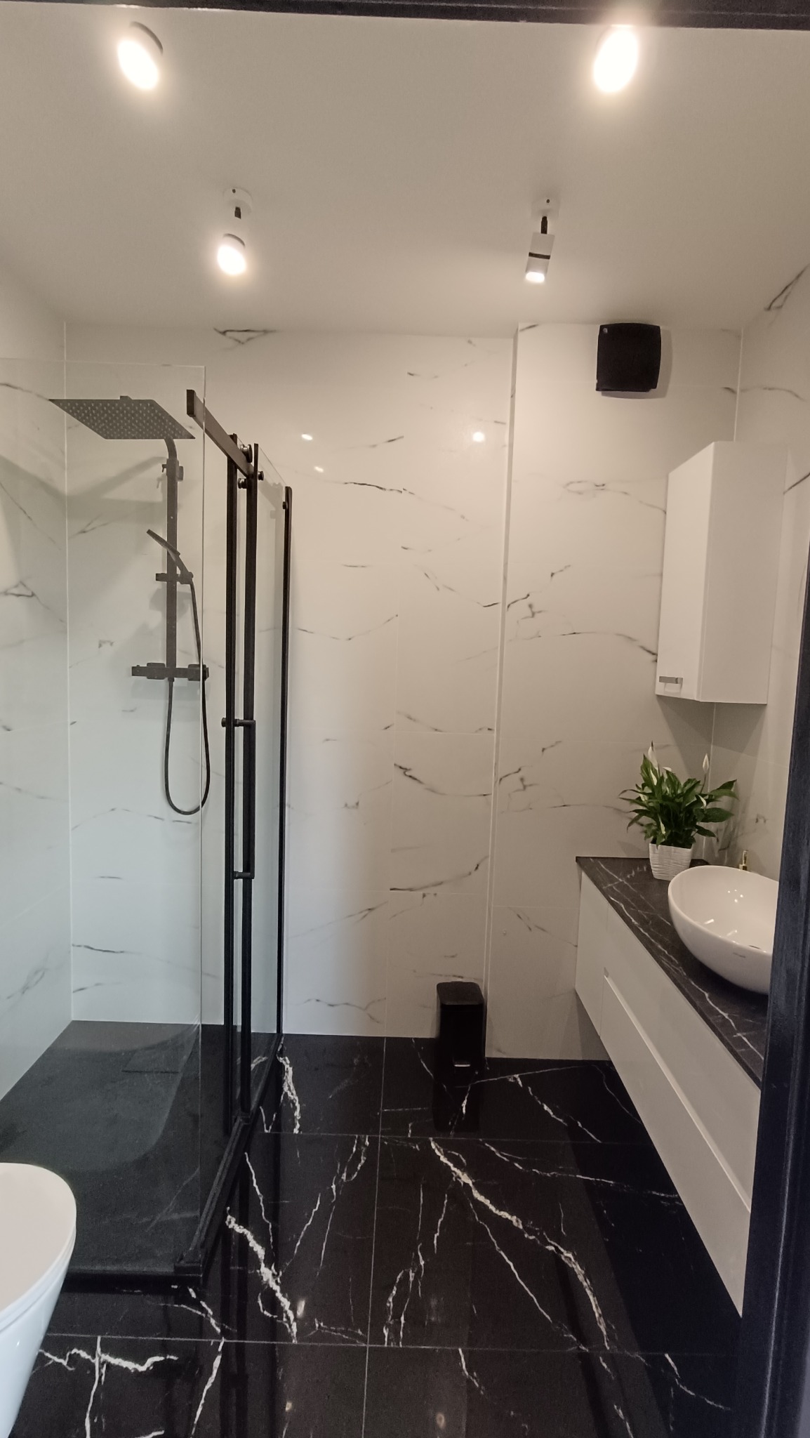 Nowoczesna łazienka z kabiną typu walk-in, wykończona płytkami imitującymi marmur w bieli i czerni, z czarnymi akcentami i oświetleniem punktowym.