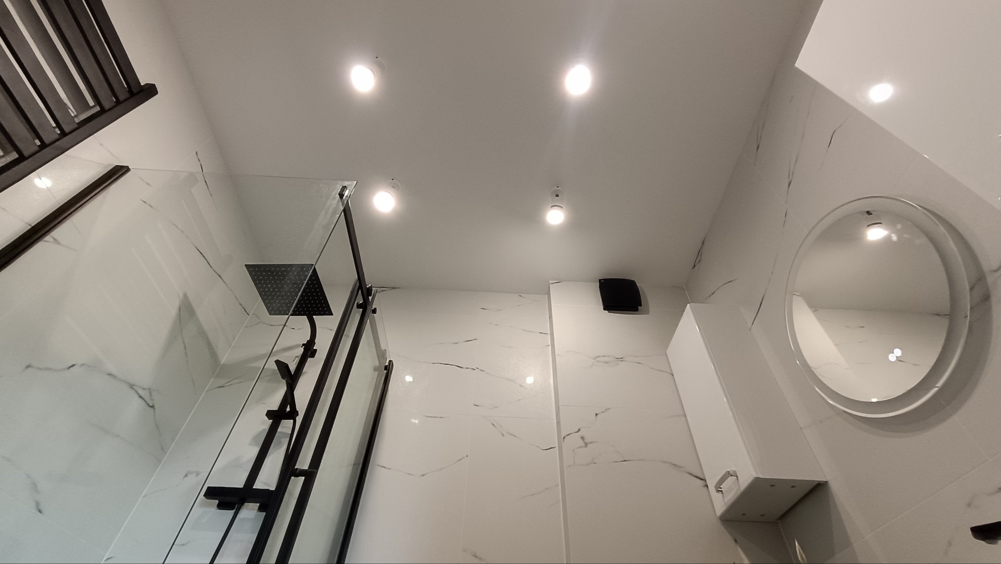 Nowoczesna łazienka z kabiną prysznicową typu walk-in, wykończona białymi płytkami imitującymi marmur. Widoczny czarny zestaw prysznicowy i okrągłe lustro.