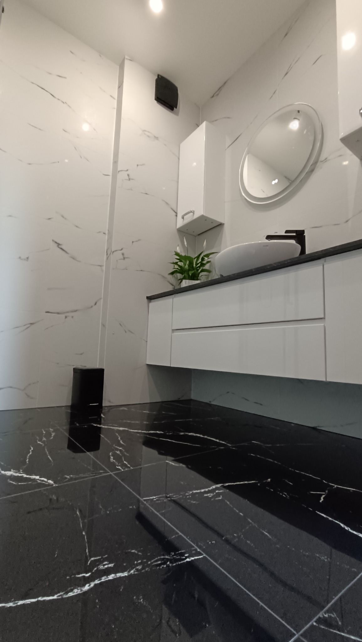 Błyszcząca, czarno-biała łazienka z owalnym lustrem, szafką pod umywalką i rośliną. Minimalistyczny design z marmurkowym wzorem na ścianach i podłodze.