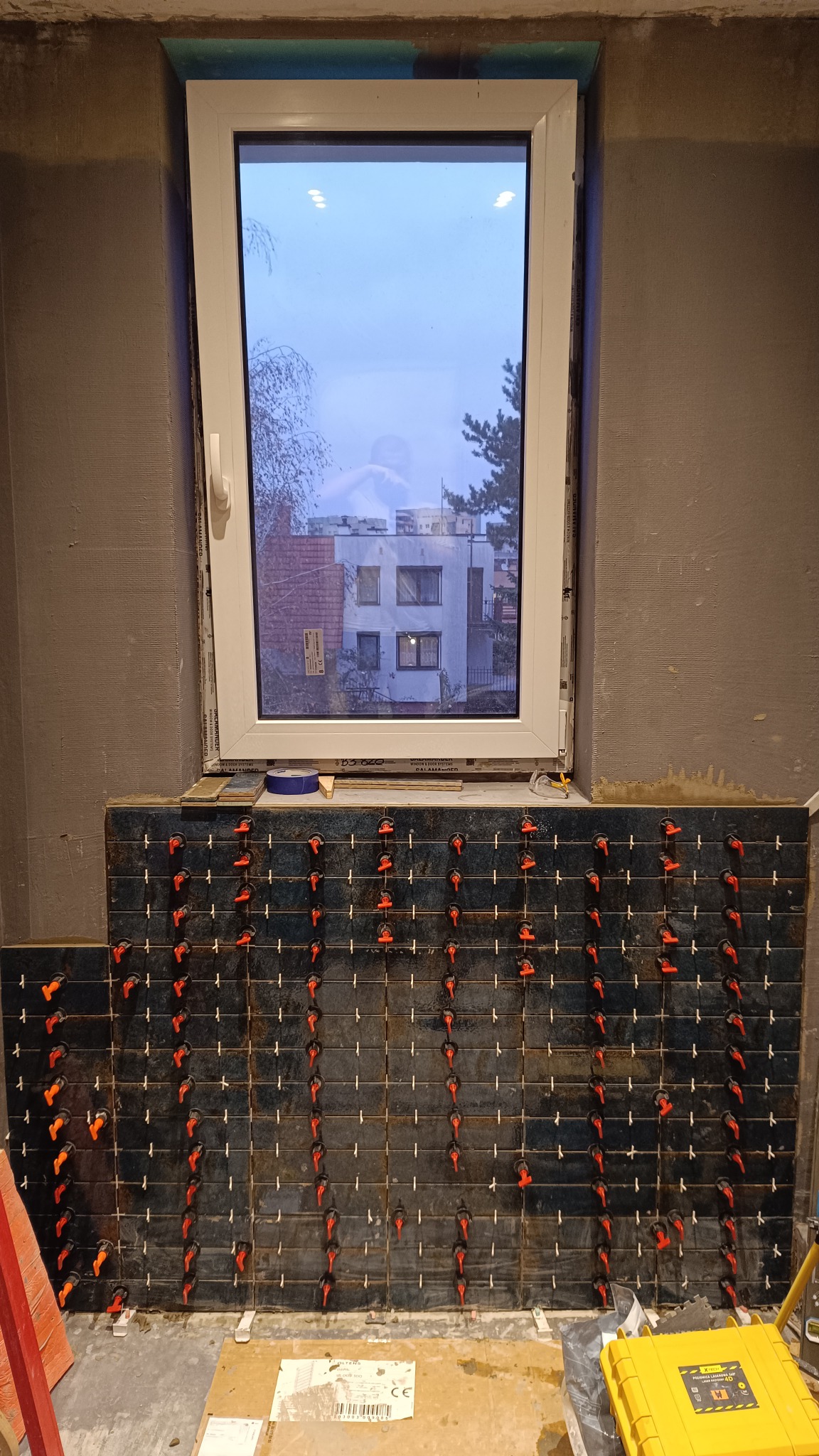 Układanie ciemnych płytek pod oknem z użyciem czerwonych klipsów poziomujących. Widoczne narzędzia glazurnicze i surowa ściana. Praca w trakcie realizacji.