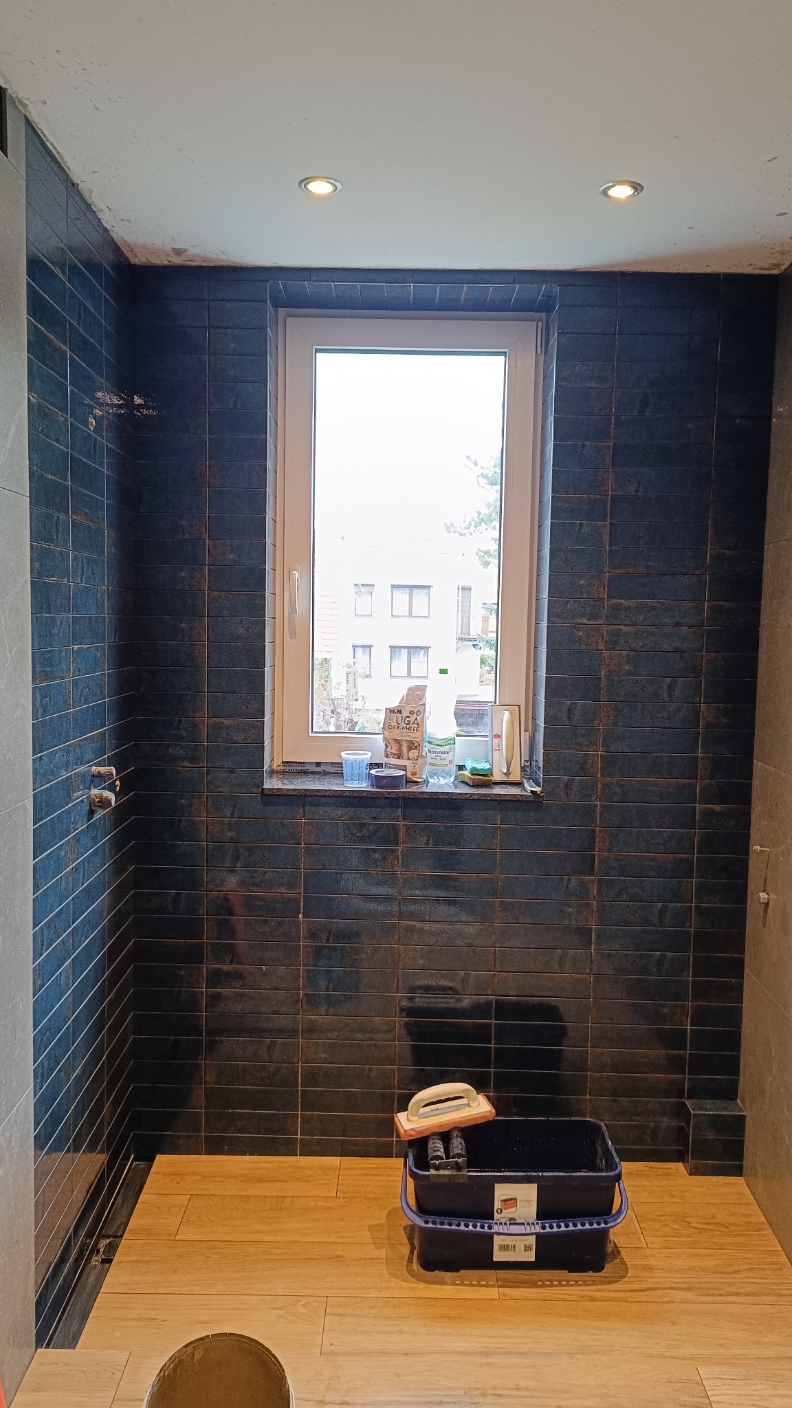 Niebieska, wąska glazura w kabinie prysznicowej z oknem. Na podłodze panele imitujące drewno. Widoczne narzędzia glazurnicze.