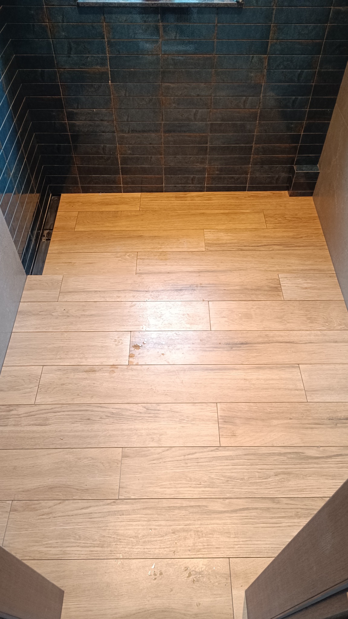 Ukończony prysznic typu walk-in z drewnopodobną podłogą i ciemnymi, błyszczącymi płytkami na ścianach. Widoczny odpływ liniowy.