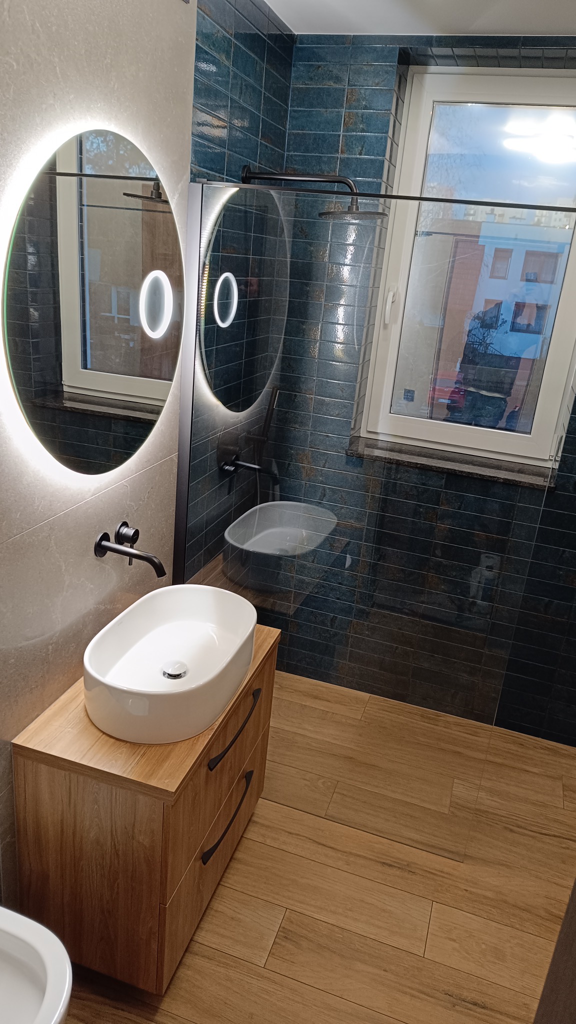 Nowoczesna łazienka z owalnym lustrem LED, umywalką nablatową na drewnianej szafce i kabiną prysznicową za szklaną ścianką. Granatowe płytki na ścianach.
