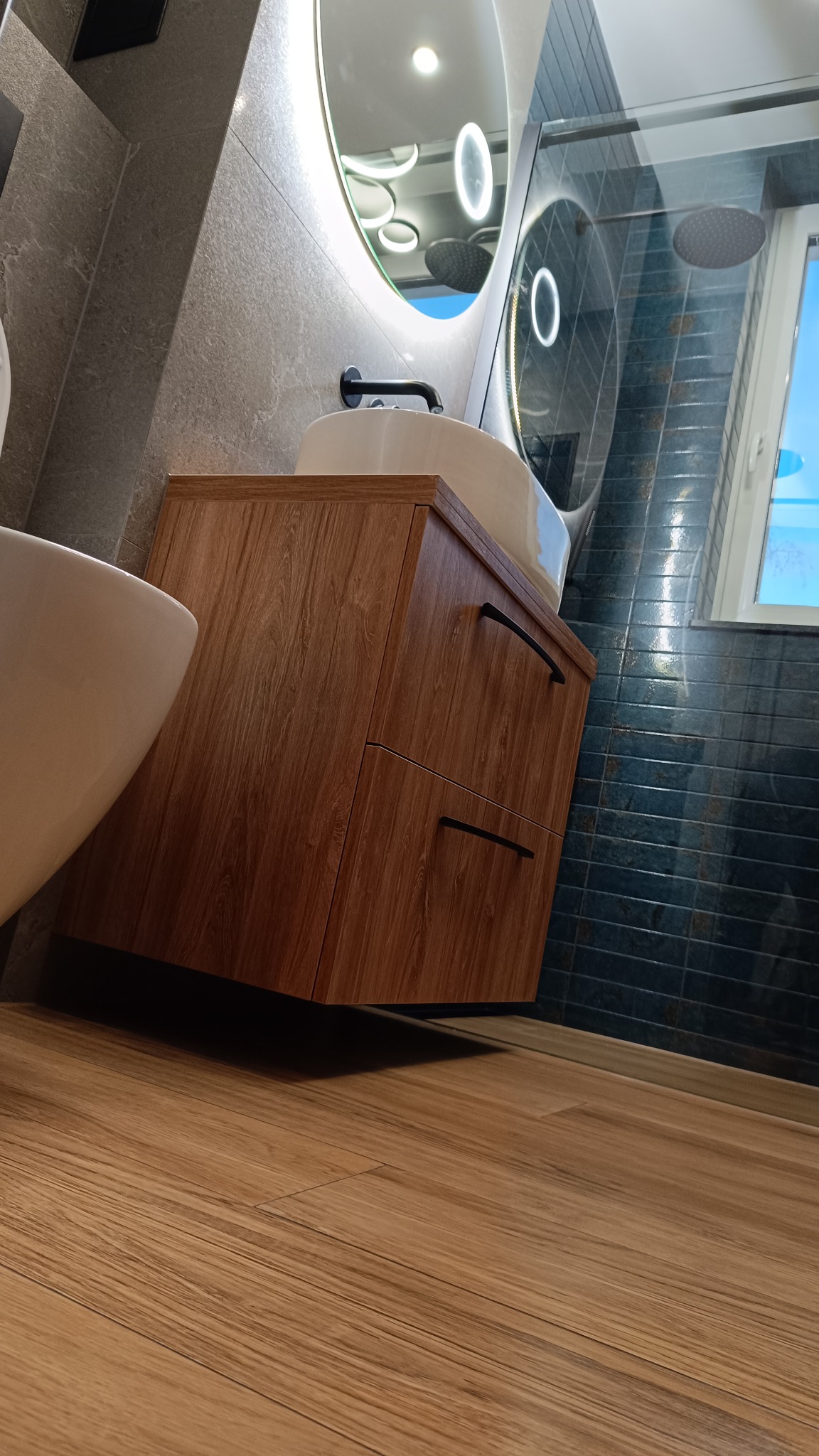 Nowoczesna łazienka z drewnianą szafką pod umywalką, okrągłym lustrem z podświetleniem LED i ciemnymi płytkami na ścianie. Widok z niskiego kąta.