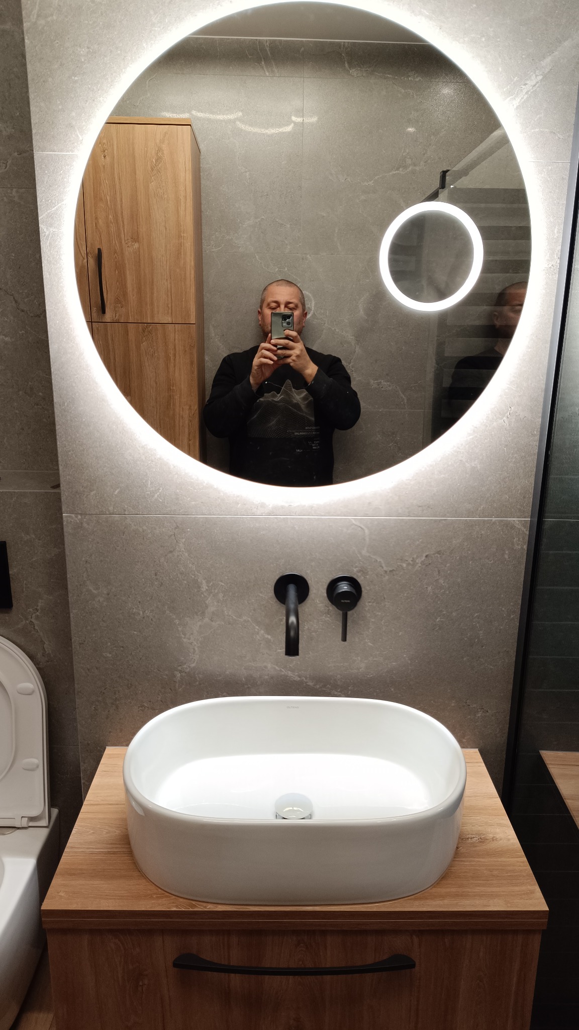 Nowoczesna łazienka z okrągłym lustrem LED, umywalką nablatową na drewnianej szafce i czarną armaturą na tle szarych płytek. Osoba robiąca zdjęcie widoczna w lustrze.