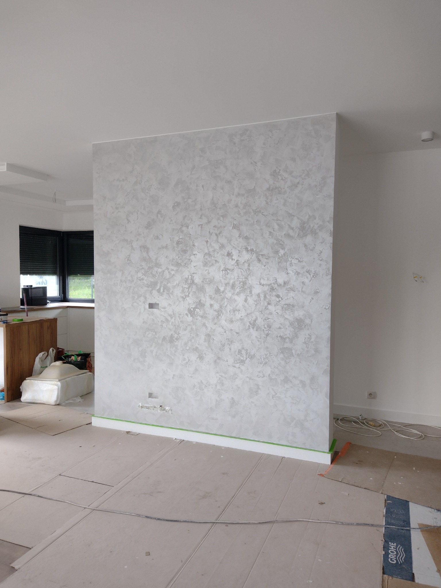 Ściana z dekoracyjnym, srebrzystym tynkiem strukturalnym. Podłoga zabezpieczona kartonem. Widoczne gniazdka elektryczne i listwy przypodłogowe. Oświetlenie sufitowe.