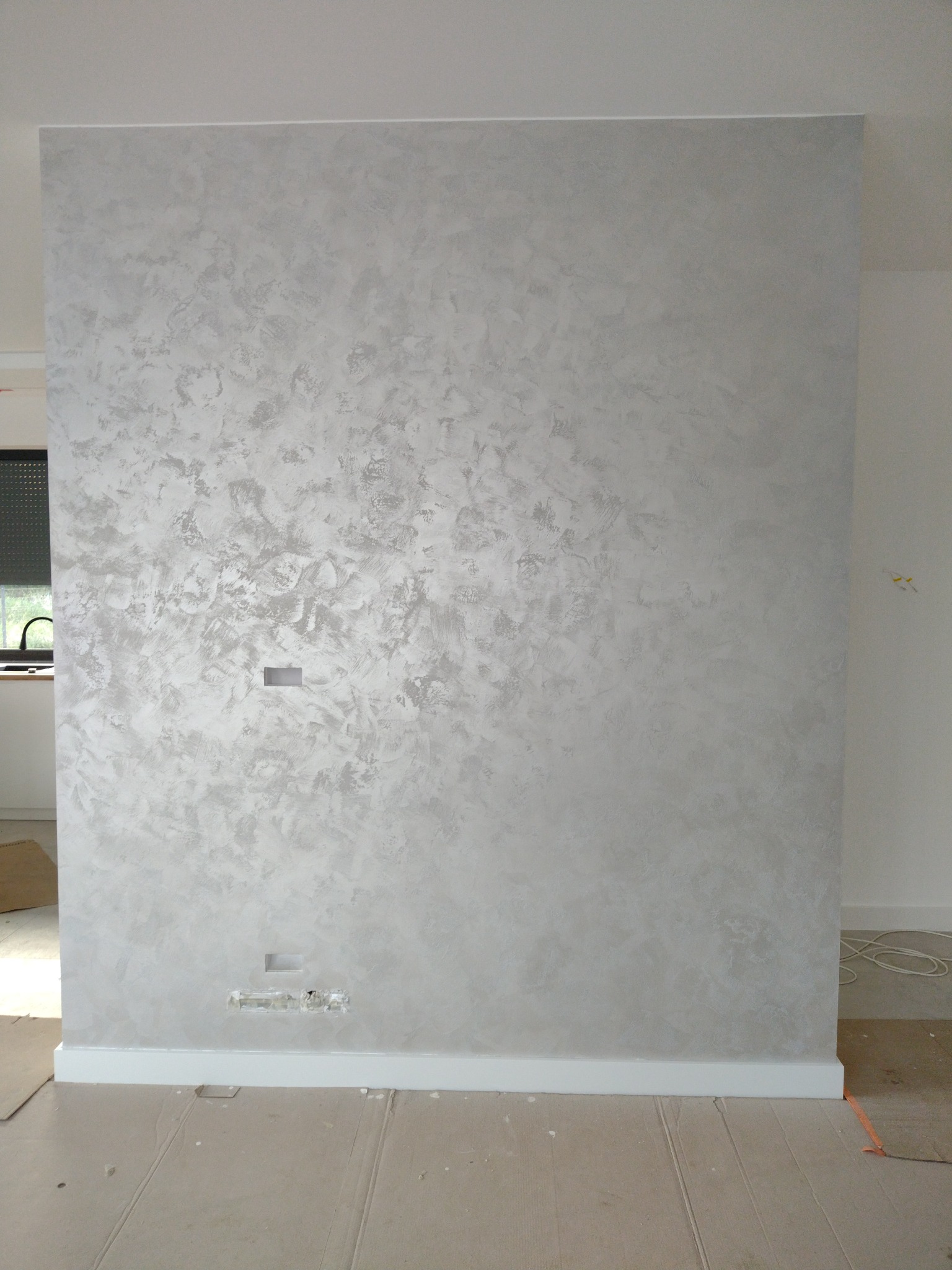 Ściana z dekoracyjną, srebrzystą fakturą w nowym wnętrzu. Widoczne gniazdka elektryczne i listwa przypodłogowa. Podłoga zabezpieczona kartonem.