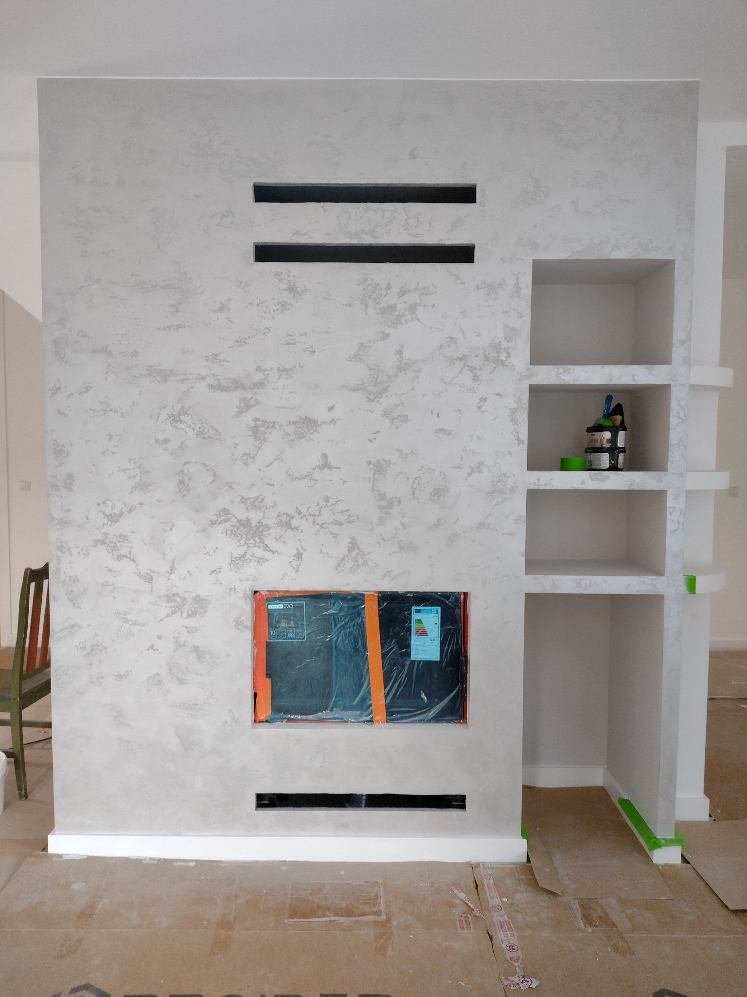 Ściana z dekoracyjnym tynkiem w odcieniach szarości, z wnękami i półkami, w trakcie wykończenia. Na podłodze kartony ochronne.