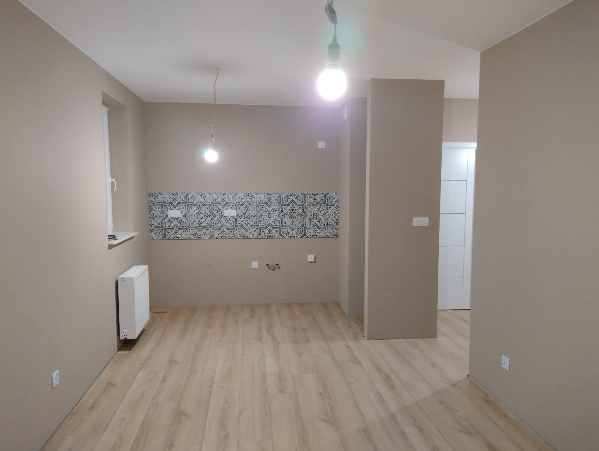 Wykończone wnętrze pokoju z panelami podłogowymi, pomalowanymi ścianami w odcieniach beżu i fragmentem ściany z dekoracyjnymi płytkami. Widoczne gniazdka elektryczne.