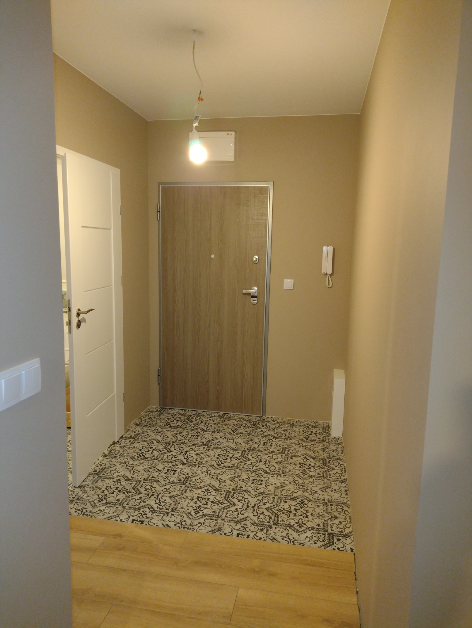 Korytarz po remoncie: beżowe ściany, drewnopodobna podłoga i wzorzysta posadzka przy drzwiach wejściowych. Widoczny biały włącznik światła i domofon.