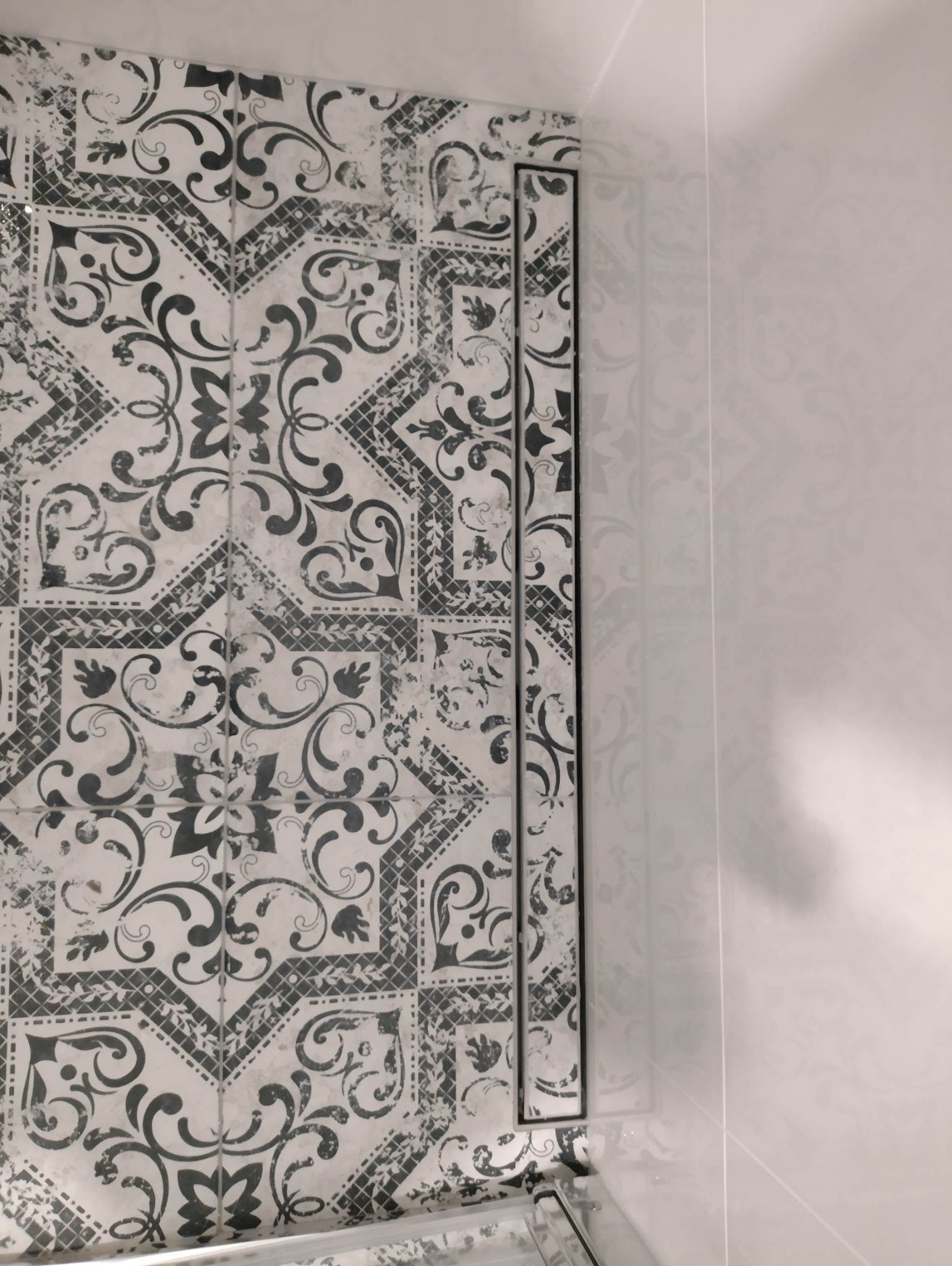 Wzorzyste, czarno-białe płytki w łazience, ułożone do rogu z minimalistycznymi białymi płytkami. Widoczny czarny profil wykończeniowy.