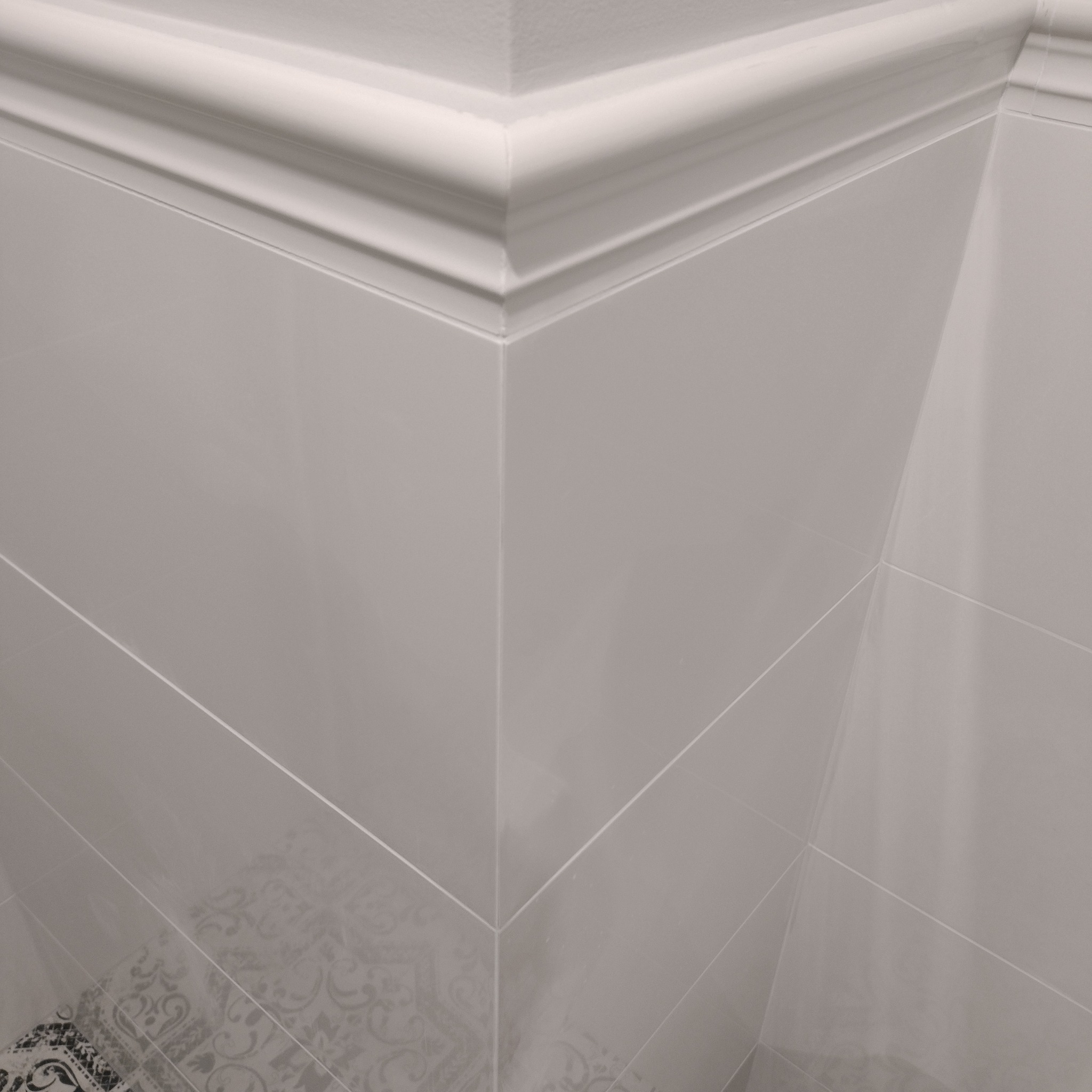 Wykończony narożnik łazienki z szarymi, błyszczącymi płytkami, białą listwą przypodłogową i fragmentem dekoracyjnej płytki podłogowej w rogu.