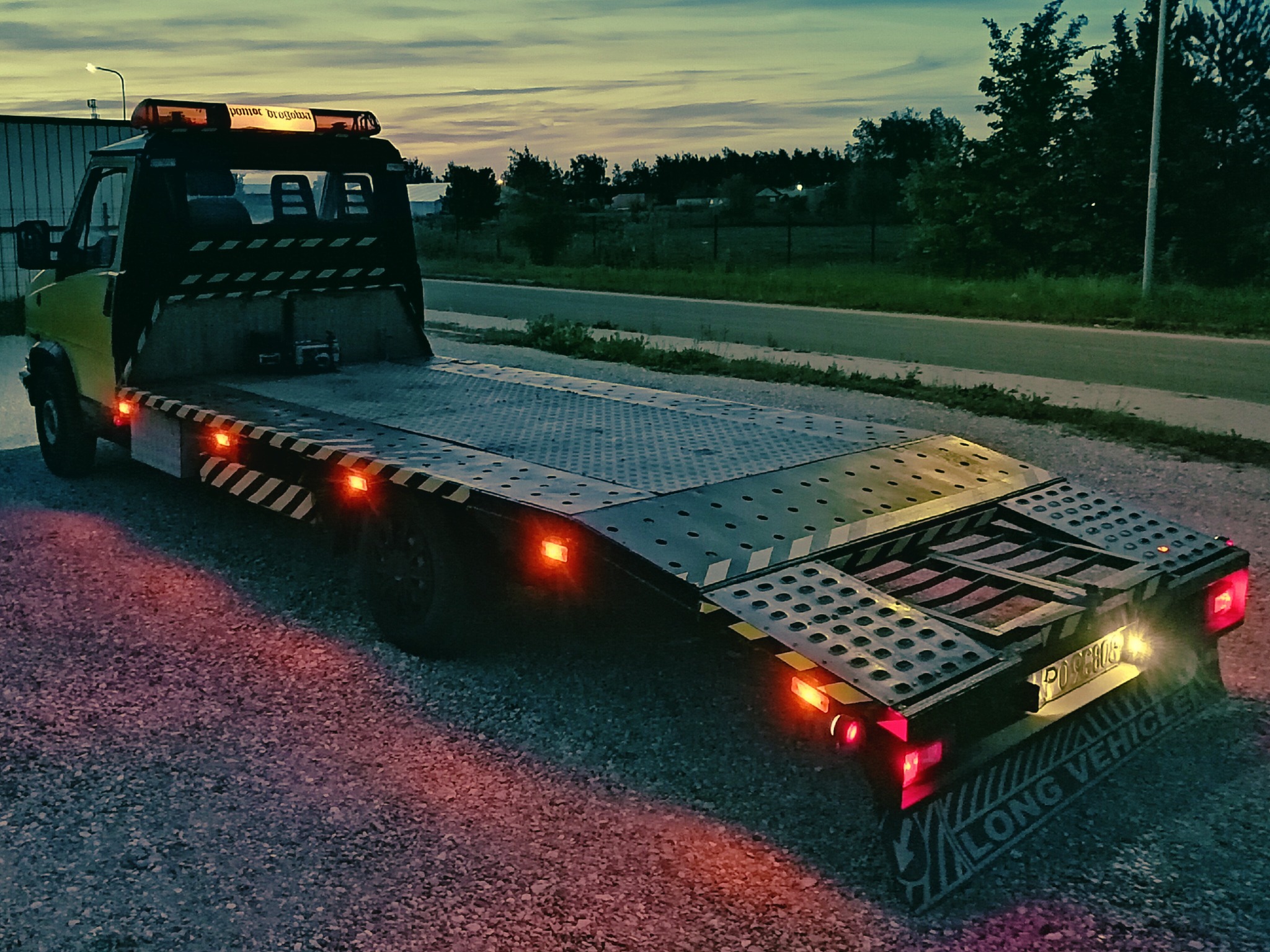 Żółta laweta z włączonymi światłami ostrzegawczymi stoi na żwirowym podłożu o zmierzchu. Widoczny napis 'LONG VEHICLE' i tablica rejestracyjna.