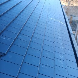 Usługi budowlane - Fragment dachu z ciemną, płaską dachówką ceramiczną w trakcie układania. Widoczne elementy konstrukcji więźby dachowej. Ujęcie z góry, lekko pod kątem.