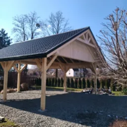 Drewniana pergola z ciemnym dachem, otoczona zielenią i wysypana kamieniami, w słoneczny dzień. Widoczne drzewa bez liści i regularnie posadzone tuje w tle.