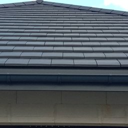 Usługi budowlane - Szczegółowy widok na ciemnoszary dach z dachówki ceramicznej, z widocznym systemem rynnowym i fragmentem ściany budynku w tle, pod lekkim kątem.