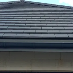 Szczegółowy widok na ciemnoszary dach z dachówki ceramicznej, z widocznym systemem rynnowym i fragmentem ściany budynku w tle, pod lekkim kątem.