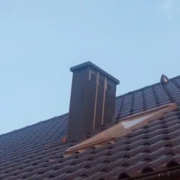 Brązowa dachówka ceramiczna z kominem wentylacyjnym i oknem dachowym w świetle dnia. Widoczny system rynnowy.