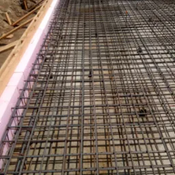 Zbrojenie pod wylewkę fundamentów z różową izolacją termiczną i drewnianym szalunkiem. Widok z góry na siatkę zbrojeniową.