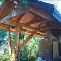 Drewniana altana z piecem murowanym i elementami kamiennymi, otoczona zielenią. Widoczna konstrukcja dachu i detale wykończenia. Ujęcie z dołu.