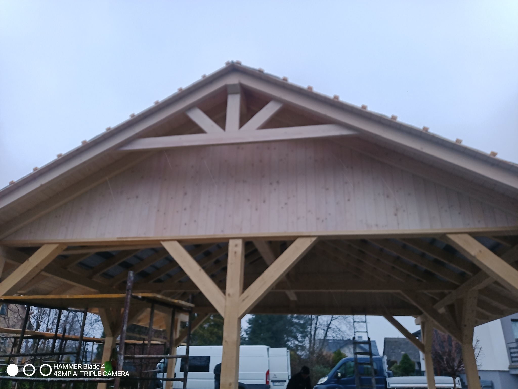 Drewniana konstrukcja altany w trakcie budowy, widoczna więźba dachowa z elementami dekoracyjnymi, surowy materiał, częściowo zadaszona, na zewnątrz pojazdy budowlane.