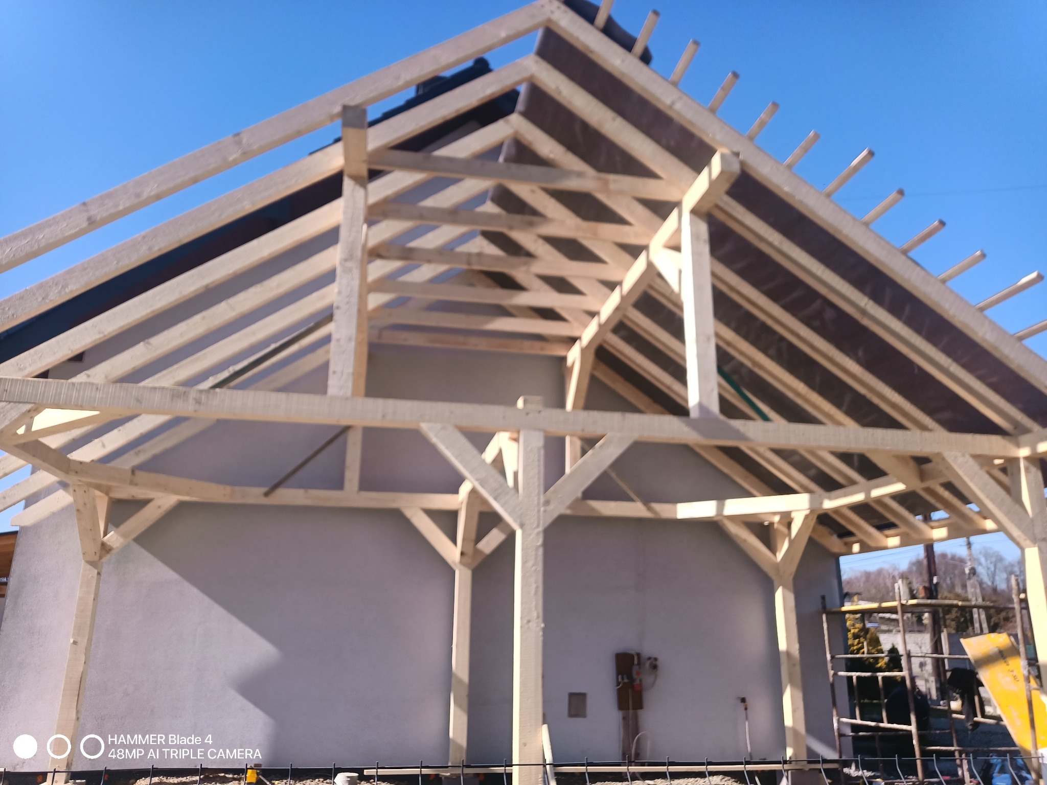 Wznoszenie drewnianej konstrukcji dachu na murowanym budynku w słoneczny dzień. Widoczna więźba dachowa i membrana dachowa. Częściowo widoczne rusztowanie.