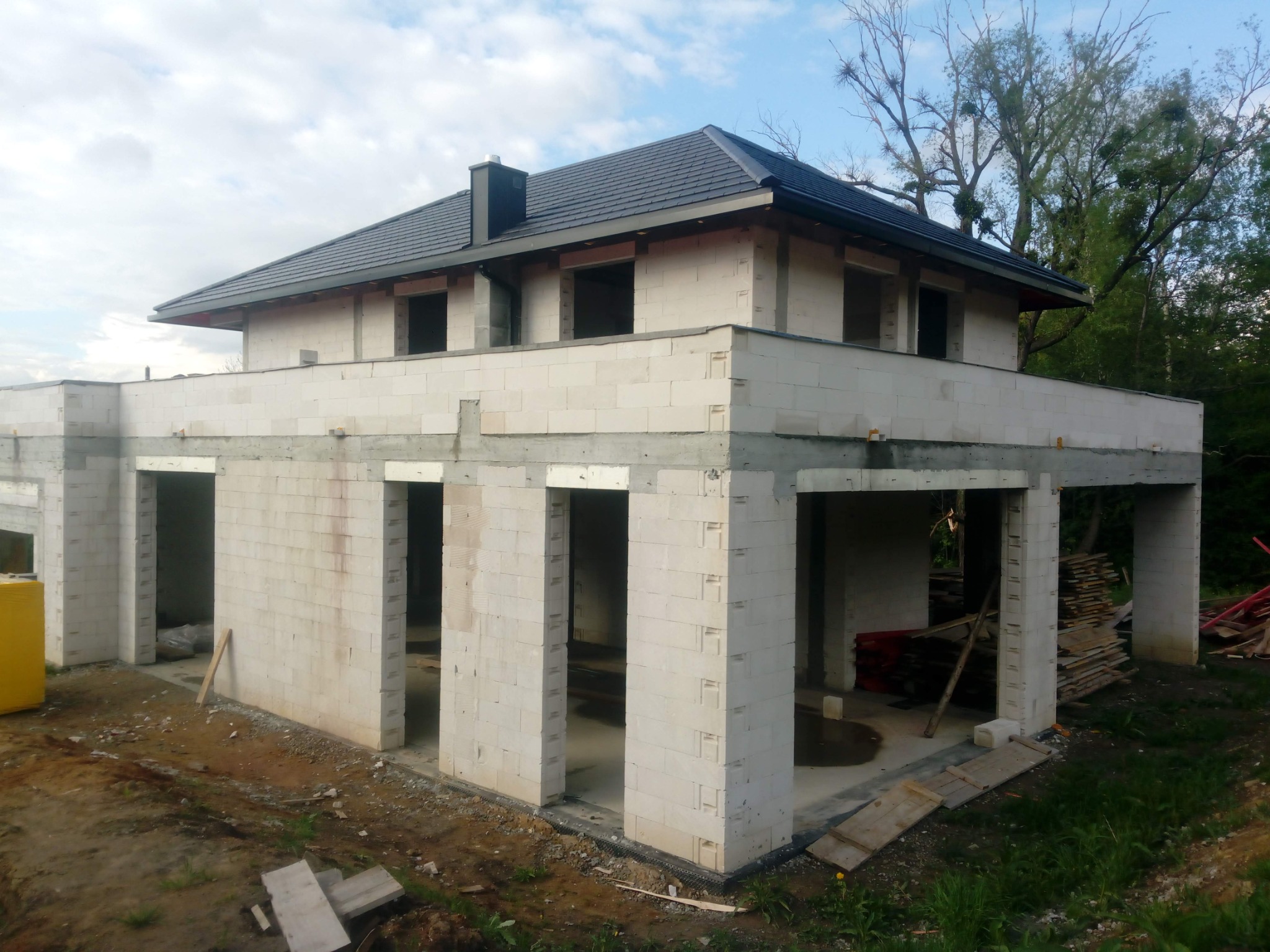 Surowy dom murowany z pustaków, z ciemnym dachem, w trakcie budowy. Widoczne niezadaszone wejścia i okna. Tło stanowi zieleń drzew i częściowo pochmurne niebo.