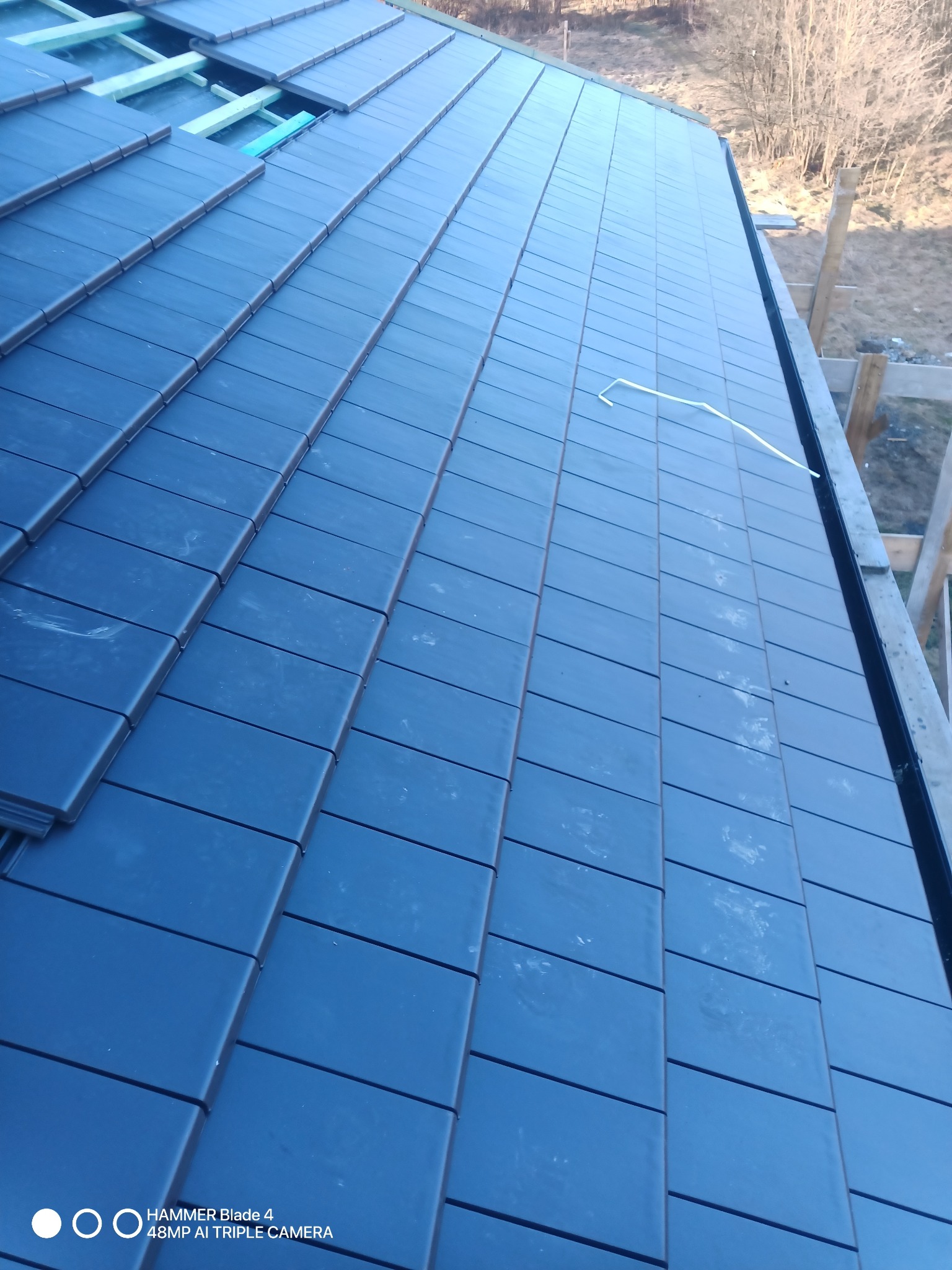 Fragment dachu z ciemną, płaską dachówką ceramiczną w trakcie układania. Widoczne elementy konstrukcji więźby dachowej. Ujęcie z góry, lekko pod kątem.