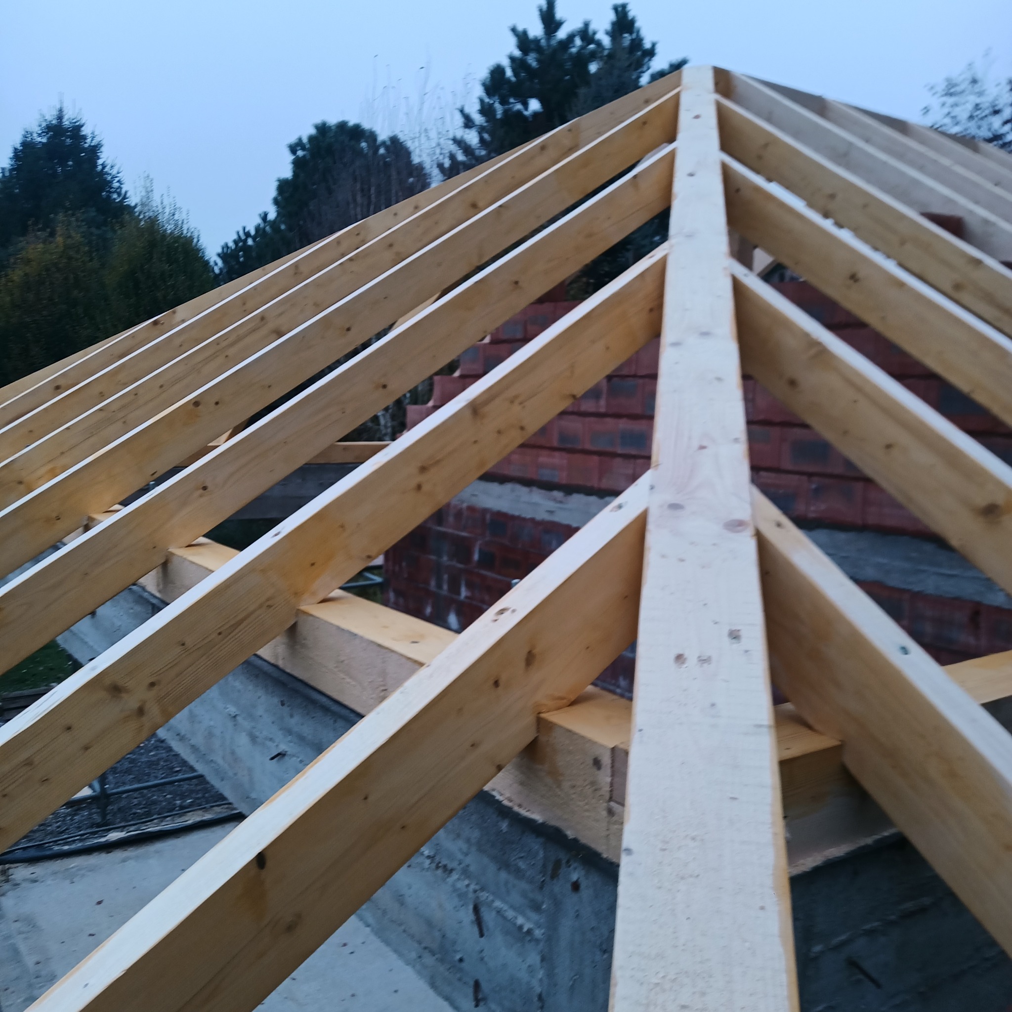 Surowa drewniana konstrukcja dachu z widoczną czerwoną cegłą w tle i zielenią drzew, uchwycona pod kątem w pochmurny dzień.