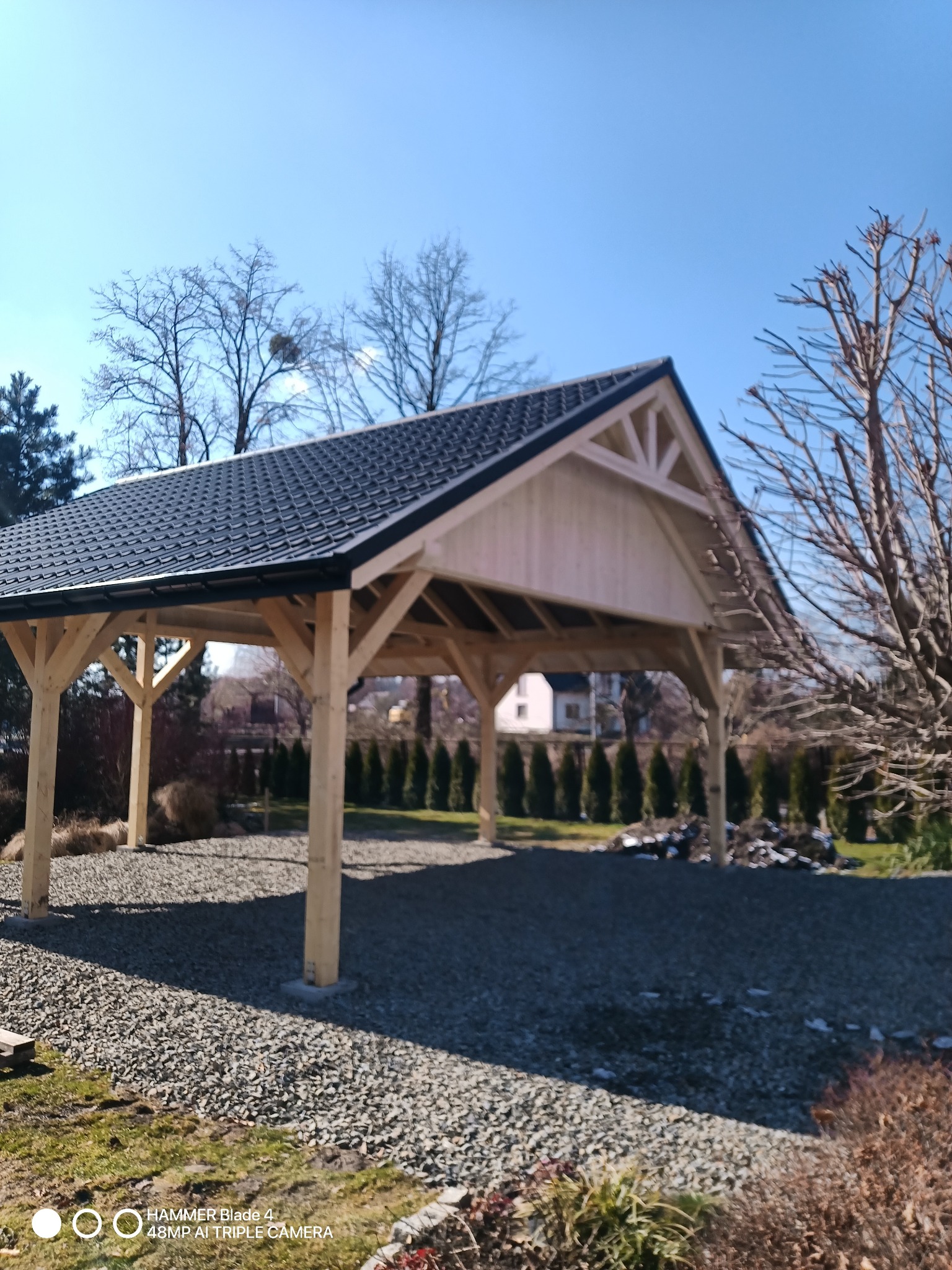 Drewniana pergola z ciemnym dachem, otoczona zielenią i wysypana kamieniami, w słoneczny dzień. Widoczne drzewa bez liści i regularnie posadzone tuje w tle.