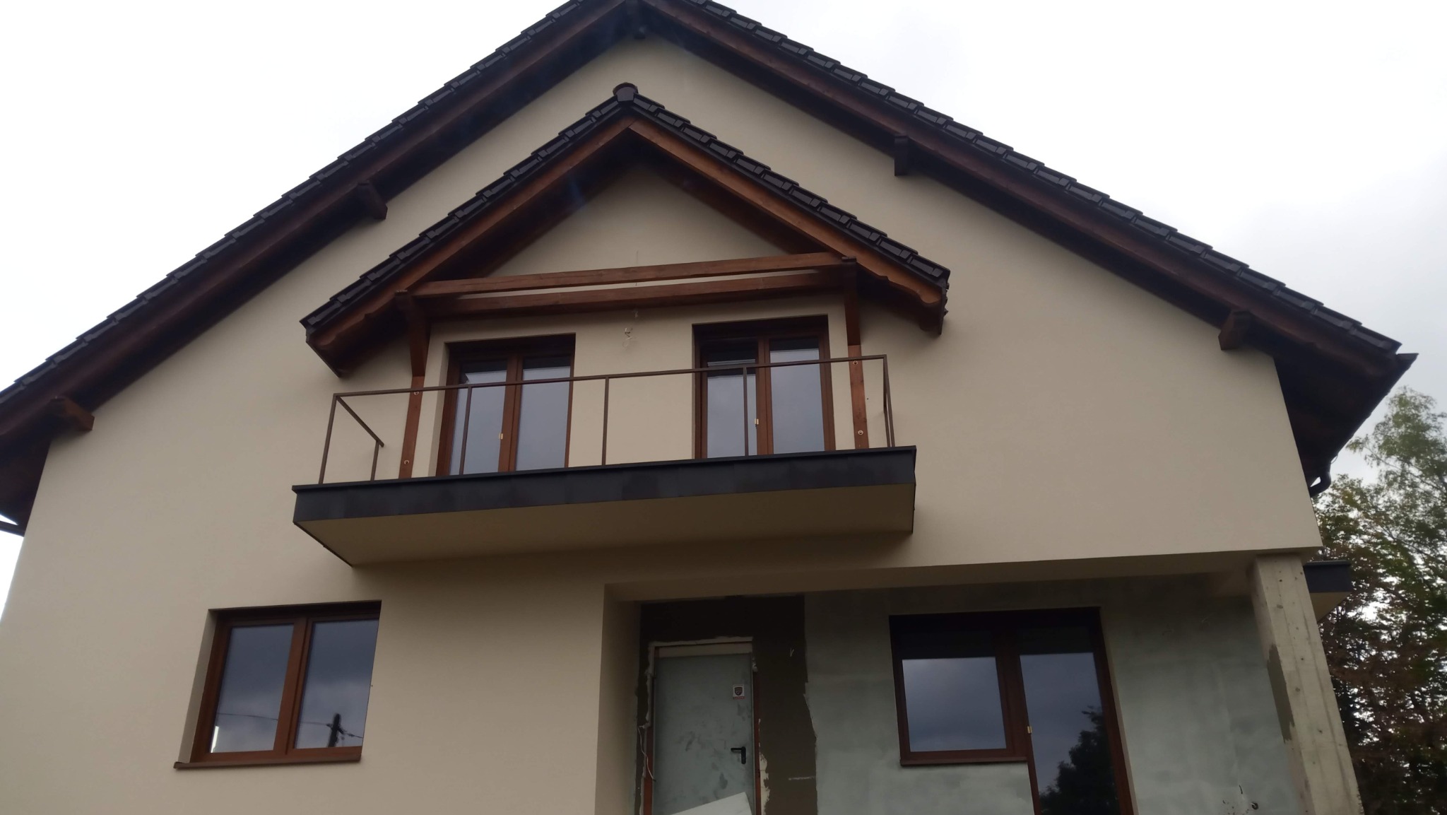 Dom z beżową elewacją, balkonem z metalową barierką i drewnianymi elementami dachu. Część elewacji w trakcie wykończenia, widoczne surowe ściany.
