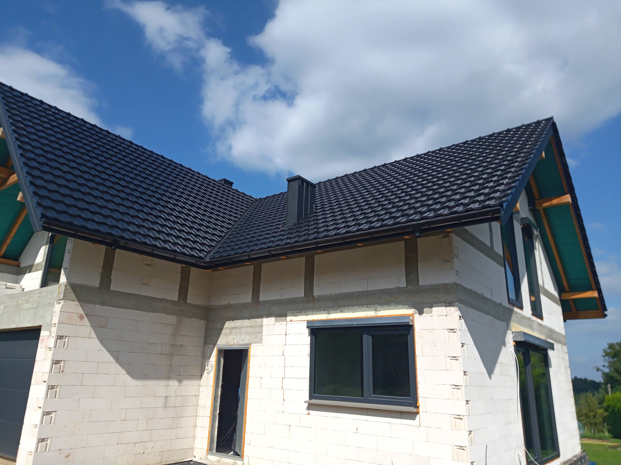 Niedokończony dom z białego pustaka, z czarnym dachem z dachówki, widok z dołu na tle błękitnego nieba z chmurami. Okna zamontowane, brak tynków.