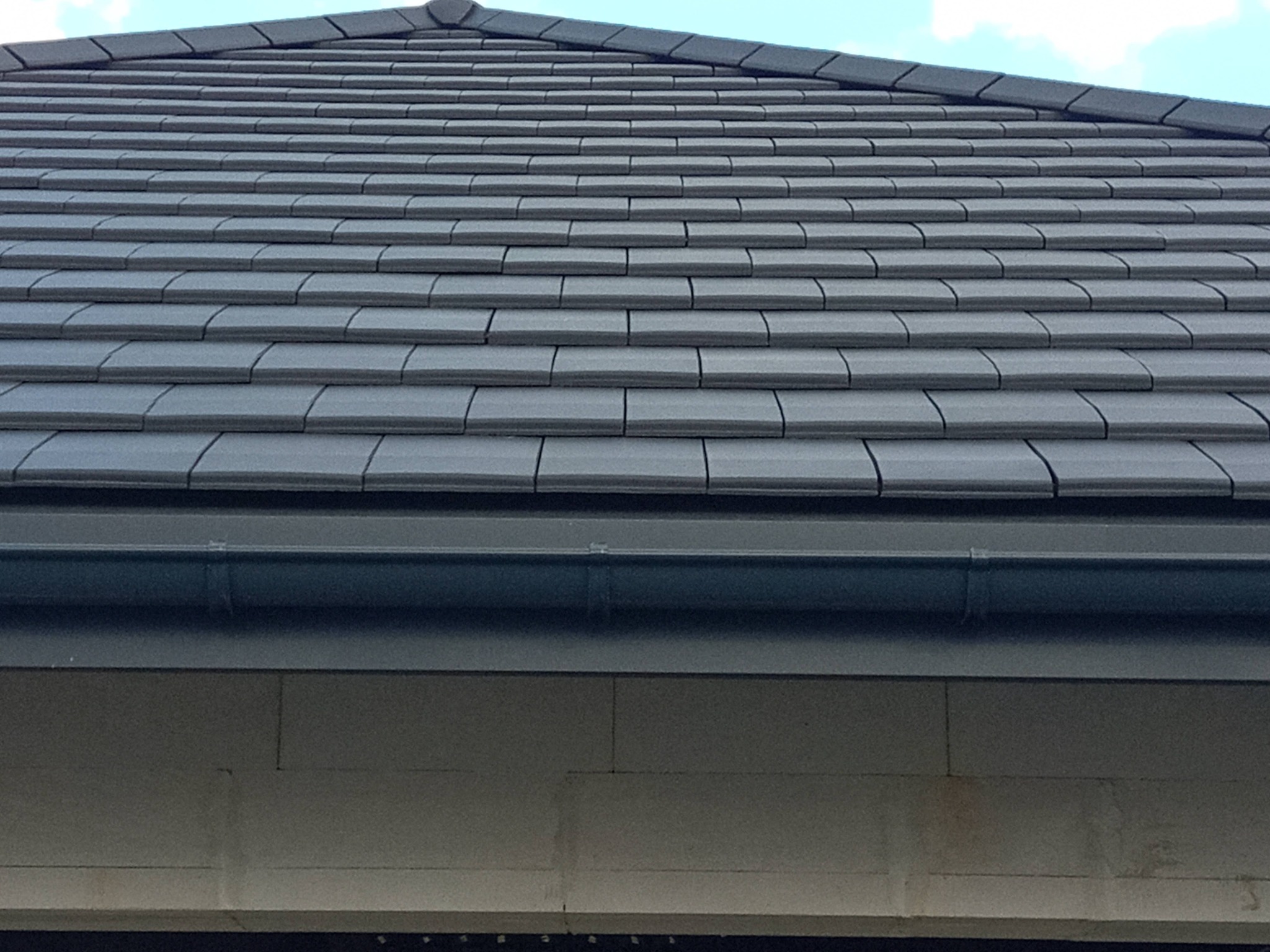 Szczegółowy widok na ciemnoszary dach z dachówki ceramicznej, z widocznym systemem rynnowym i fragmentem ściany budynku w tle, pod lekkim kątem.