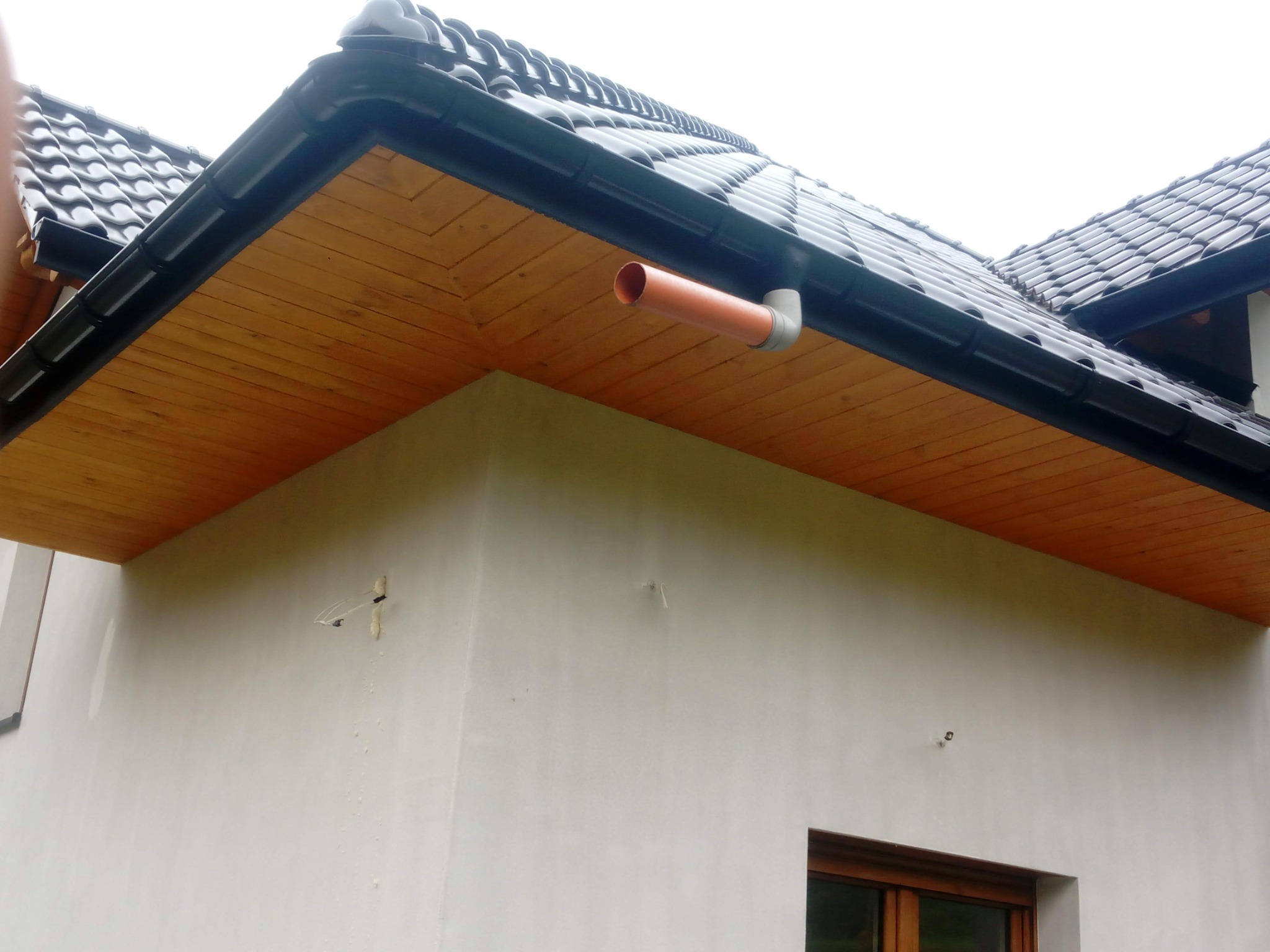 Fragment domu z ciemną dachówką, drewnianą podbitką i rynną. Detal orynnowania z rurą spustową. Ściana budynku w jasnym kolorze, widoczne okno.
