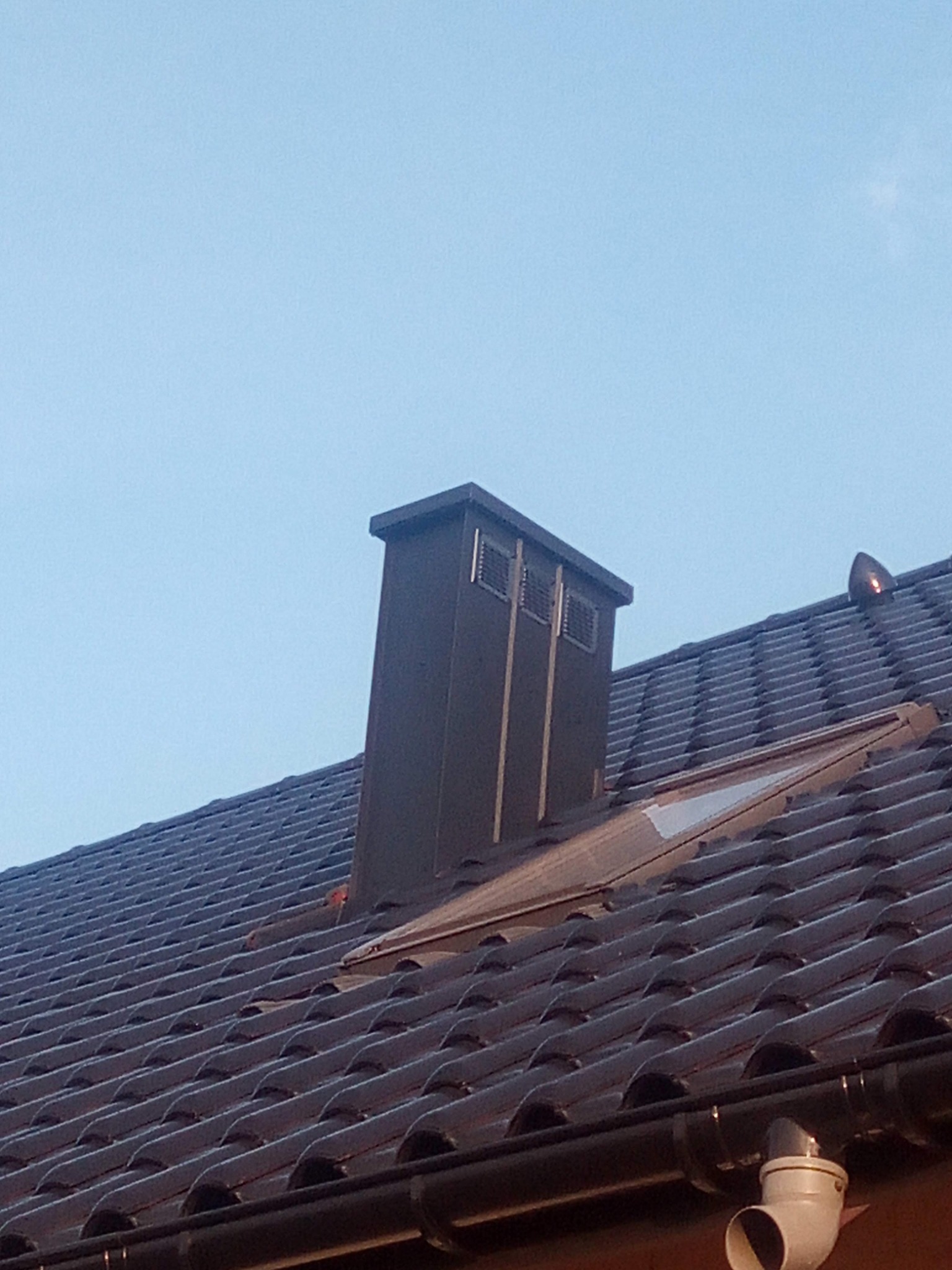 Brązowa dachówka ceramiczna z kominem wentylacyjnym i oknem dachowym w świetle dnia. Widoczny system rynnowy.