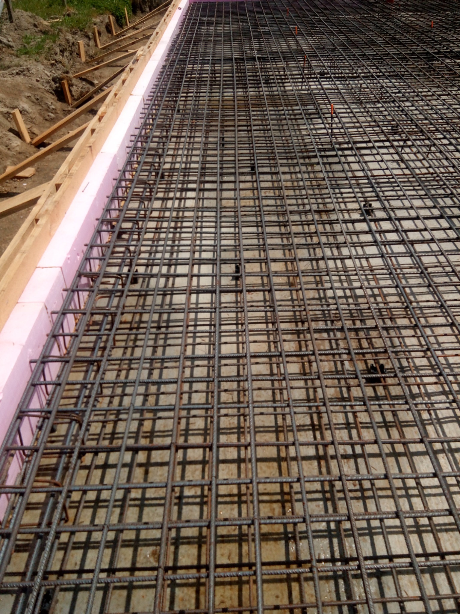 Zbrojenie pod wylewkę fundamentów z różową izolacją termiczną i drewnianym szalunkiem. Widok z góry na siatkę zbrojeniową.
