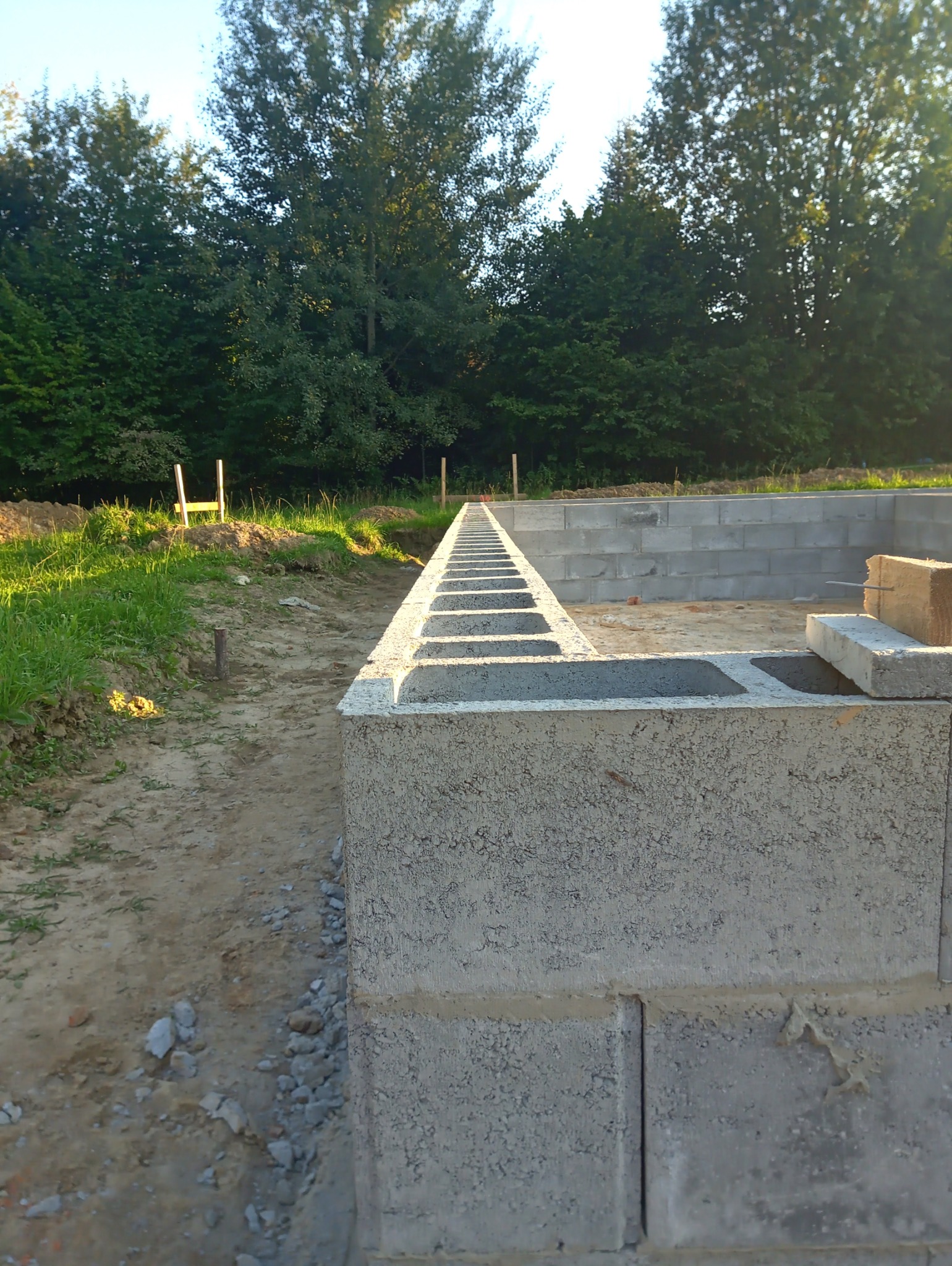 Fundamenty z bloczków betonowych w trakcie budowy domu, widok z poziomu gruntu, zieleń w tle. Elementy szalunku widoczne w oddali.