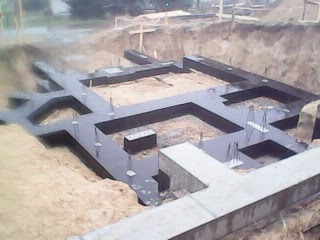 Zalane fundamenty domu w wykopie, widoczne betonowe ławy i izolacja pozioma. Etap budowy w surowym stanie otwartym, teren piaszczysty.