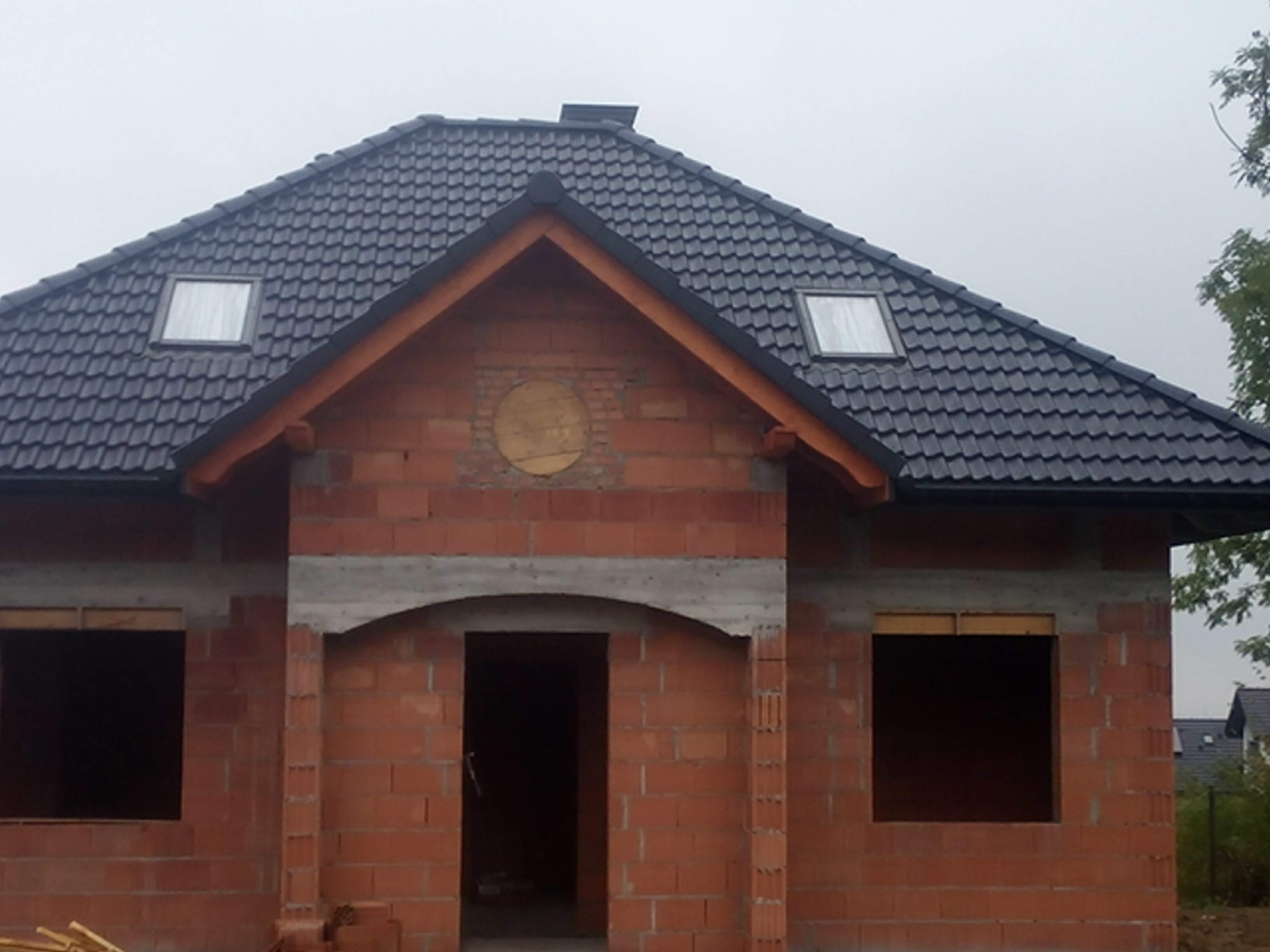 Niedokończony dom z czerwonej cegły z ciemnym dachem i oknami dachowymi. Widoczny stan surowy, brak tynków. Jesienne, pochmurne niebo w tle.