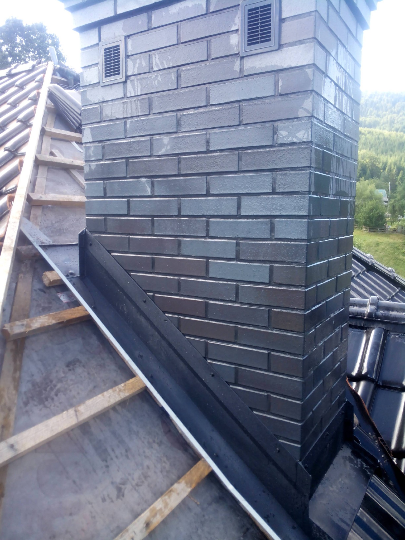 Murowany komin z odpowietrznikami, obróbka blacharska na dachu z czarnej dachówki. Widoczna więźba dachowa i zielone wzgórza w tle. Komin po deszczu.