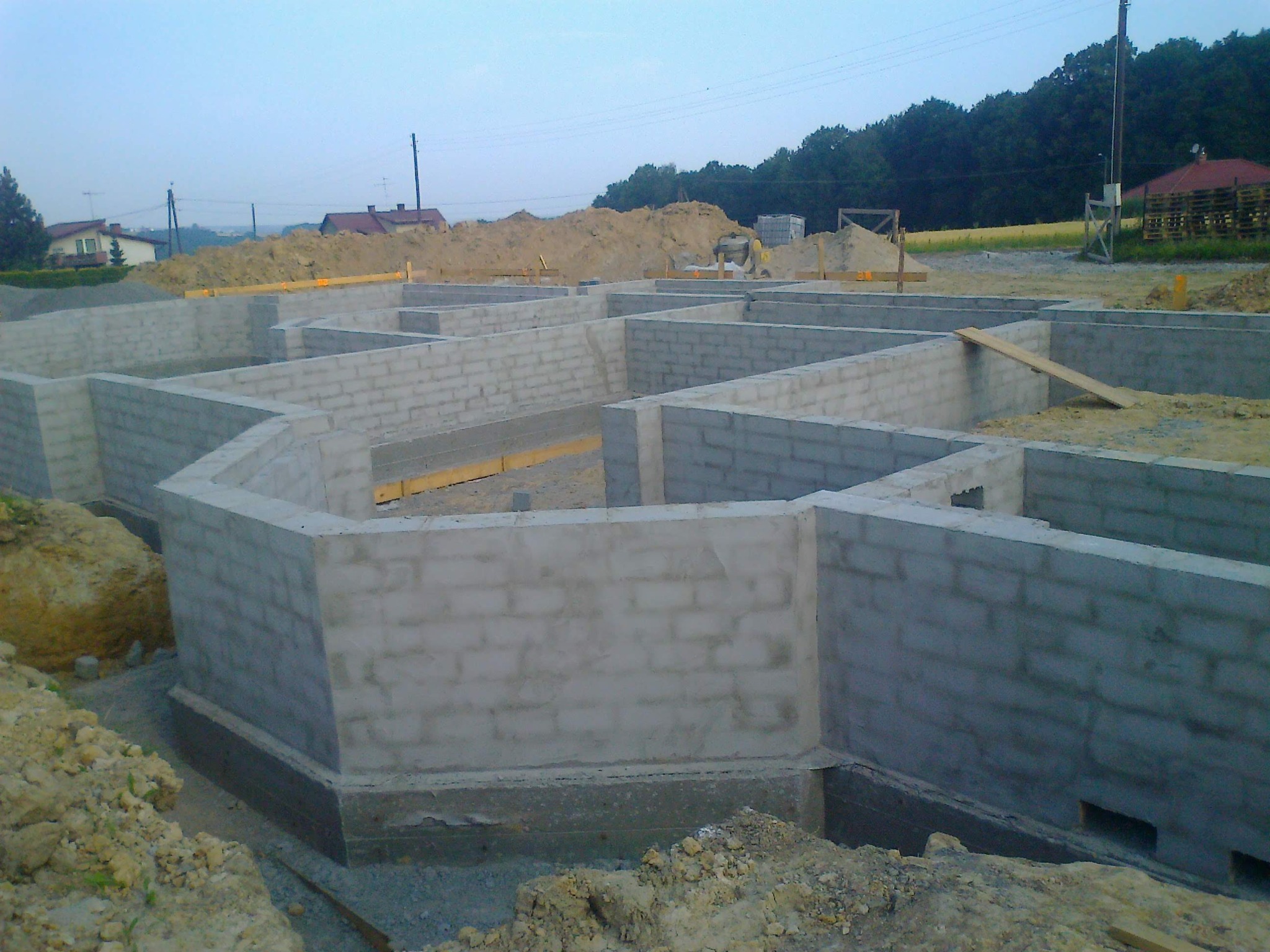 Fundamenty domu z bloczków betonowych w trakcie budowy, widok z góry. W tle hałda ziemi i zabudowania.