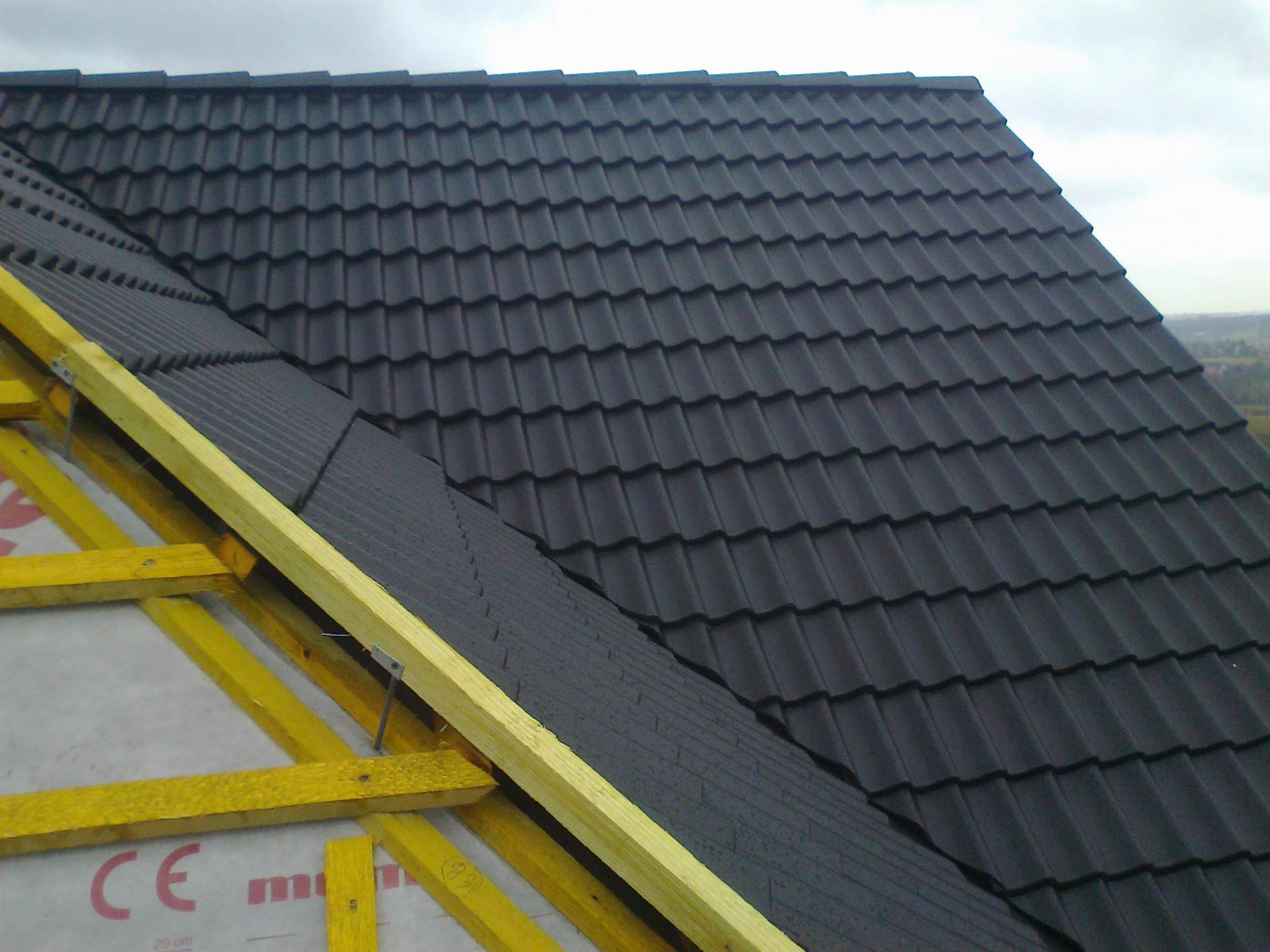 Częściowy widok dachu z czarną dachówką na drewnianej więźbie, z widoczną membraną dachową. Ujęcie z bliska, z perspektywy od dołu.