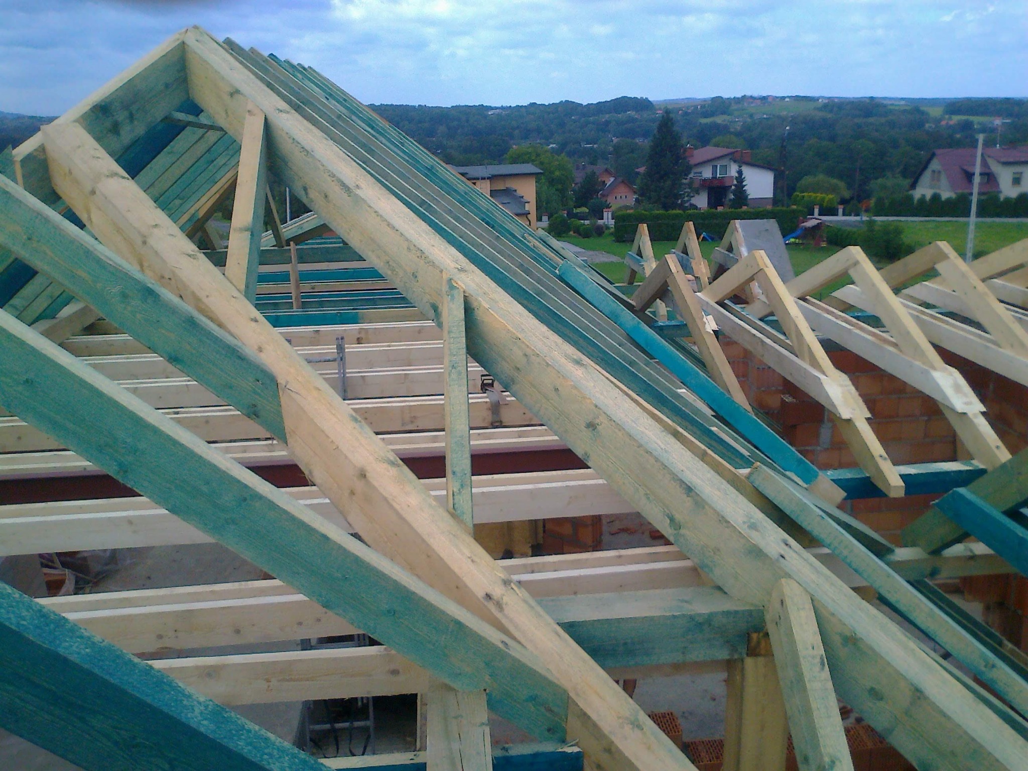 W trakcie budowy: surowa więźba dachowa z impregnowanego drewna, widok z góry na konstrukcję z elementami muru i krajobrazem w tle. Niebo pochmurne.