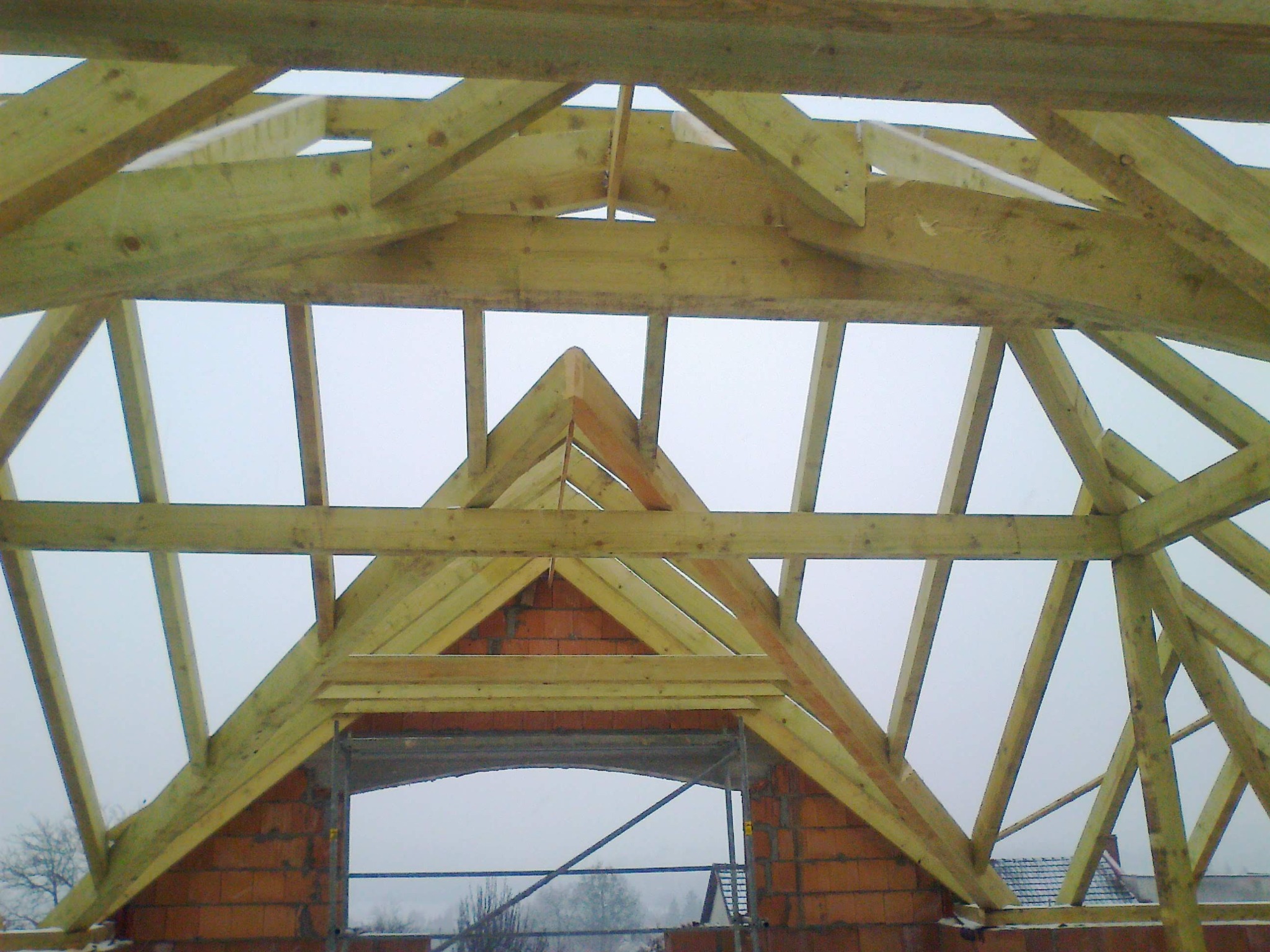 Wnętrze domu w trakcie budowy. Widoczna drewniana konstrukcja więźby dachowej z elementami murowanymi. W tle szare niebo.