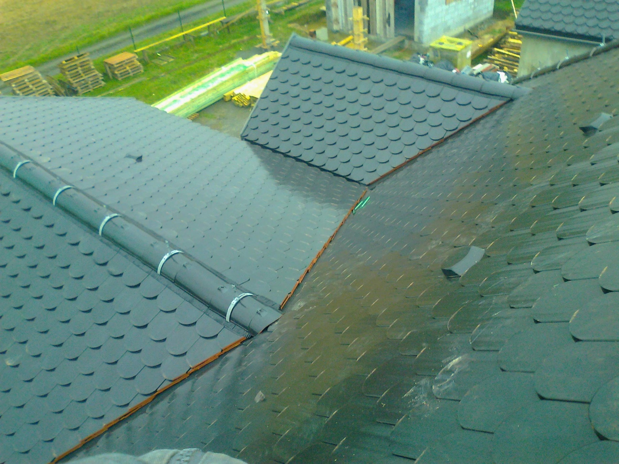 Fragment dachu pokrytego ciemną dachówką bitumiczną w kształcie rybiej łuski, z widocznymi elementami orynnowania i fragmentem budynku w tle.