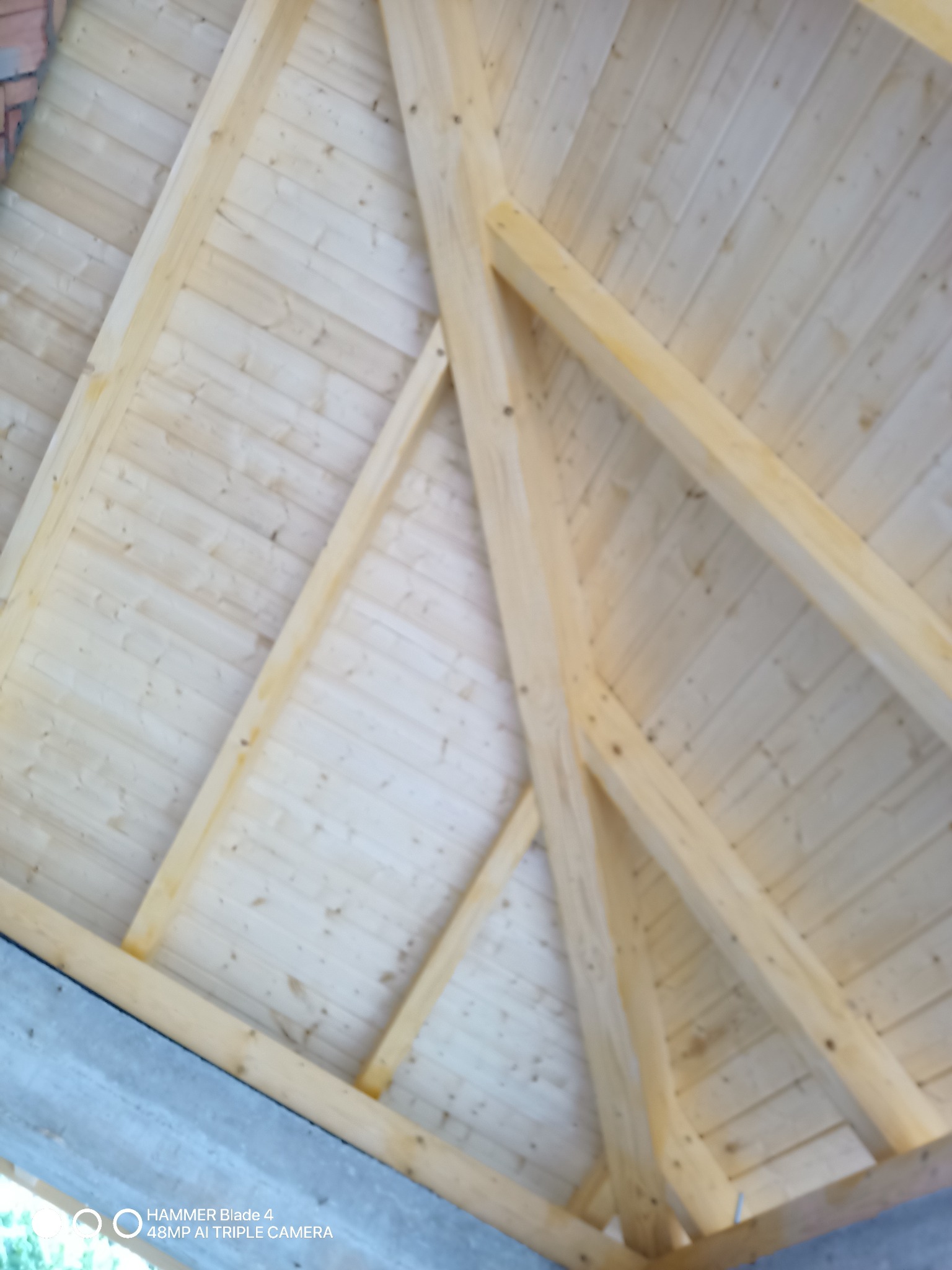 Jasna drewniana konstrukcja dachu z widocznymi belkami i deskami, wykonana pod kątem. Elementy złączone na gwoździe. Widok od dołu.