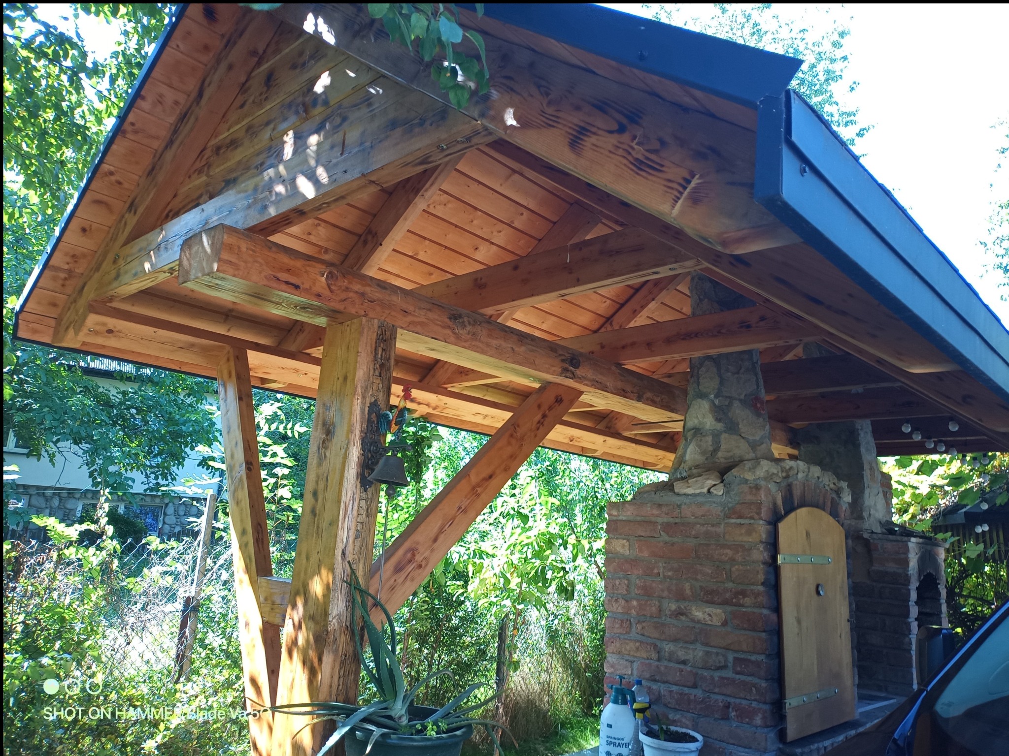 Drewniana altana z piecem murowanym i elementami kamiennymi, otoczona zielenią. Widoczna konstrukcja dachu i detale wykończenia. Ujęcie z dołu.