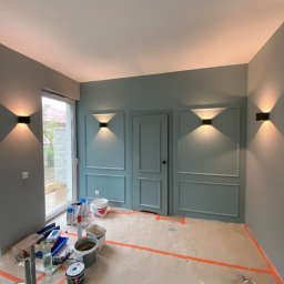 Stepan Malitskyi Standem Bud - Pokój po malowaniu w odcieniach szarości i błękitu z dekoracyjnymi listwami i nowoczesnym oświetleniem ściennym. Widoczne narzędzia malarskie.